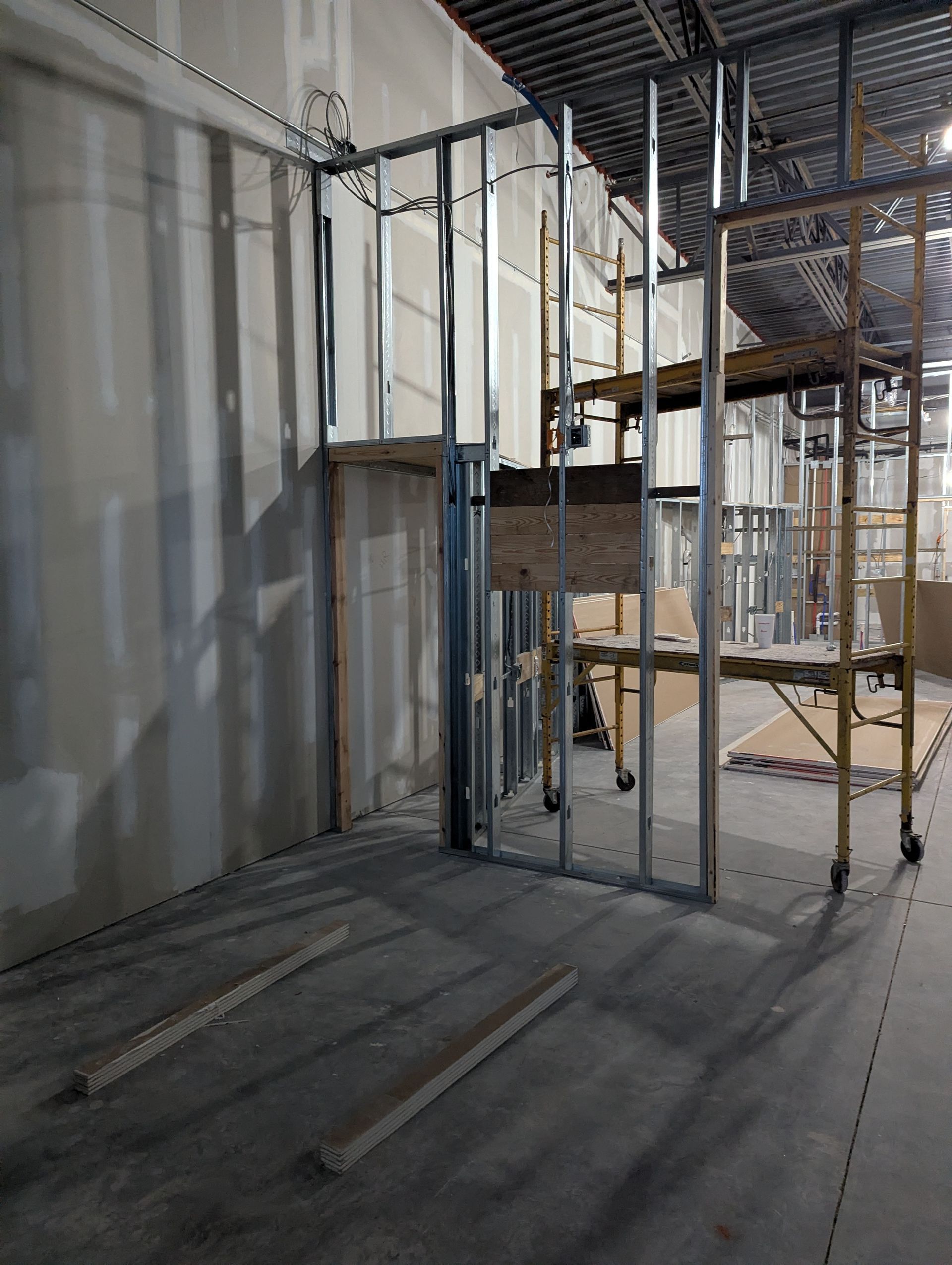 Interior view of a construction site showing metal wall studs, drywall, and yellow scaffolding on a concrete floor.