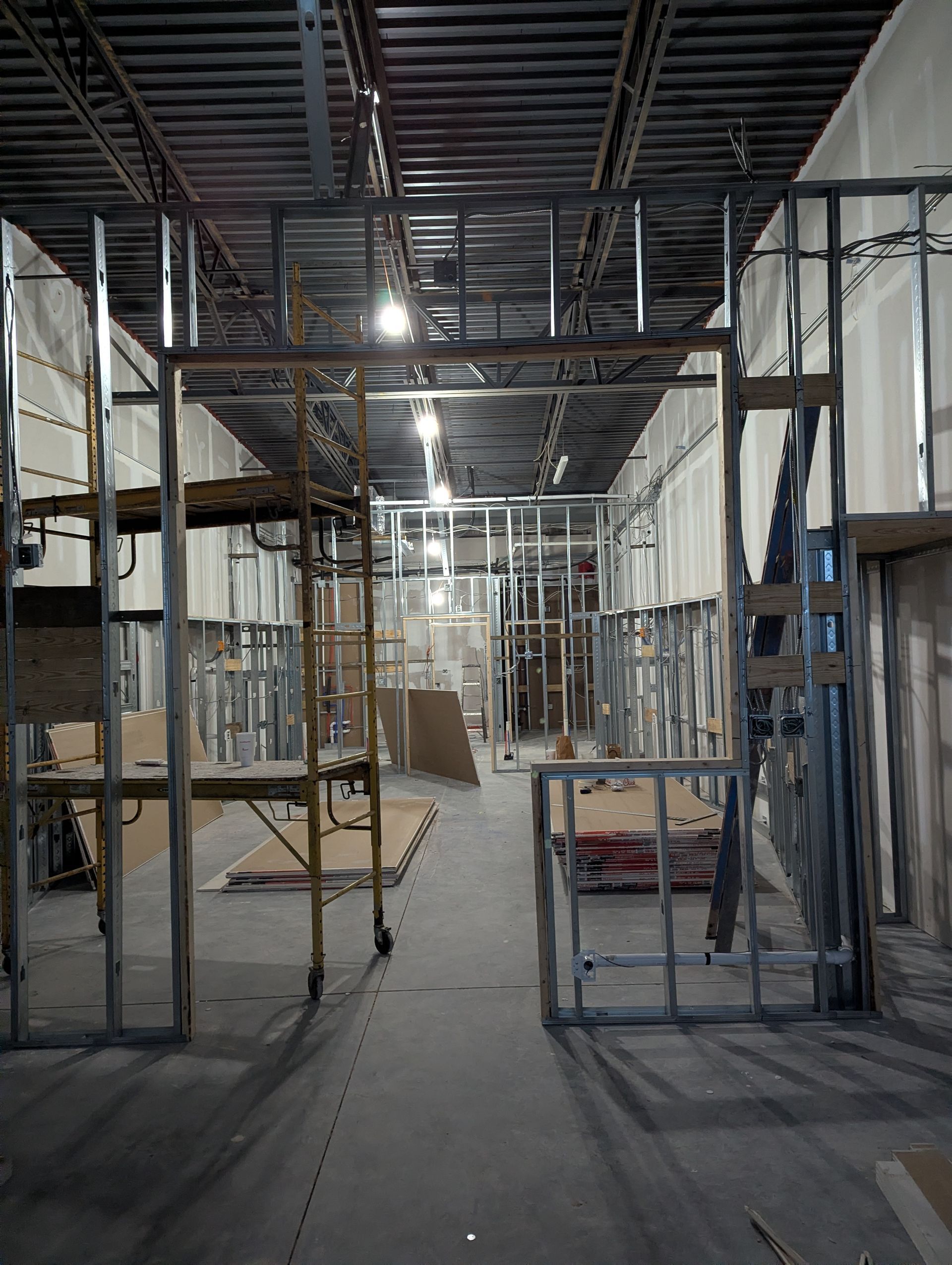 Interior view of a commercial construction site showing metal wall framing, open ceiling rafters, and scaffolding.