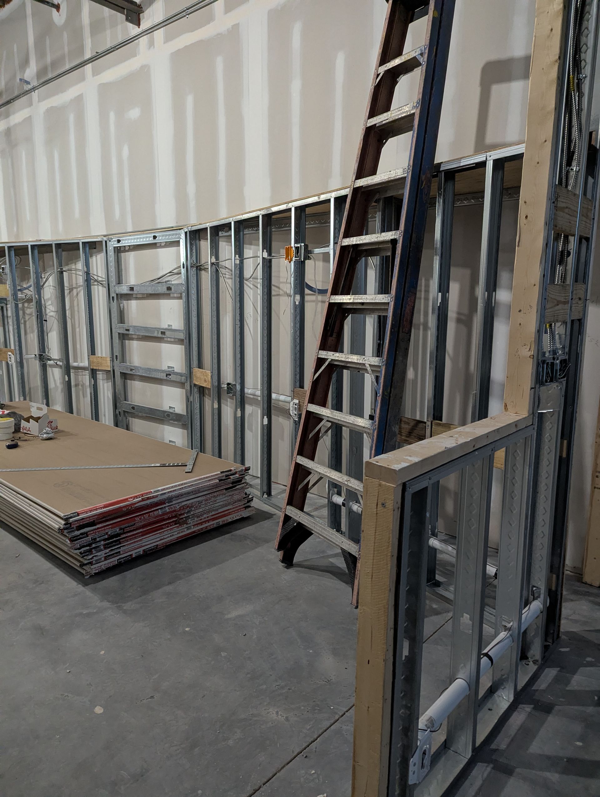 A construction site showing metal wall studs, a ladder leaning against the wall, and a stack of drywall sheets.