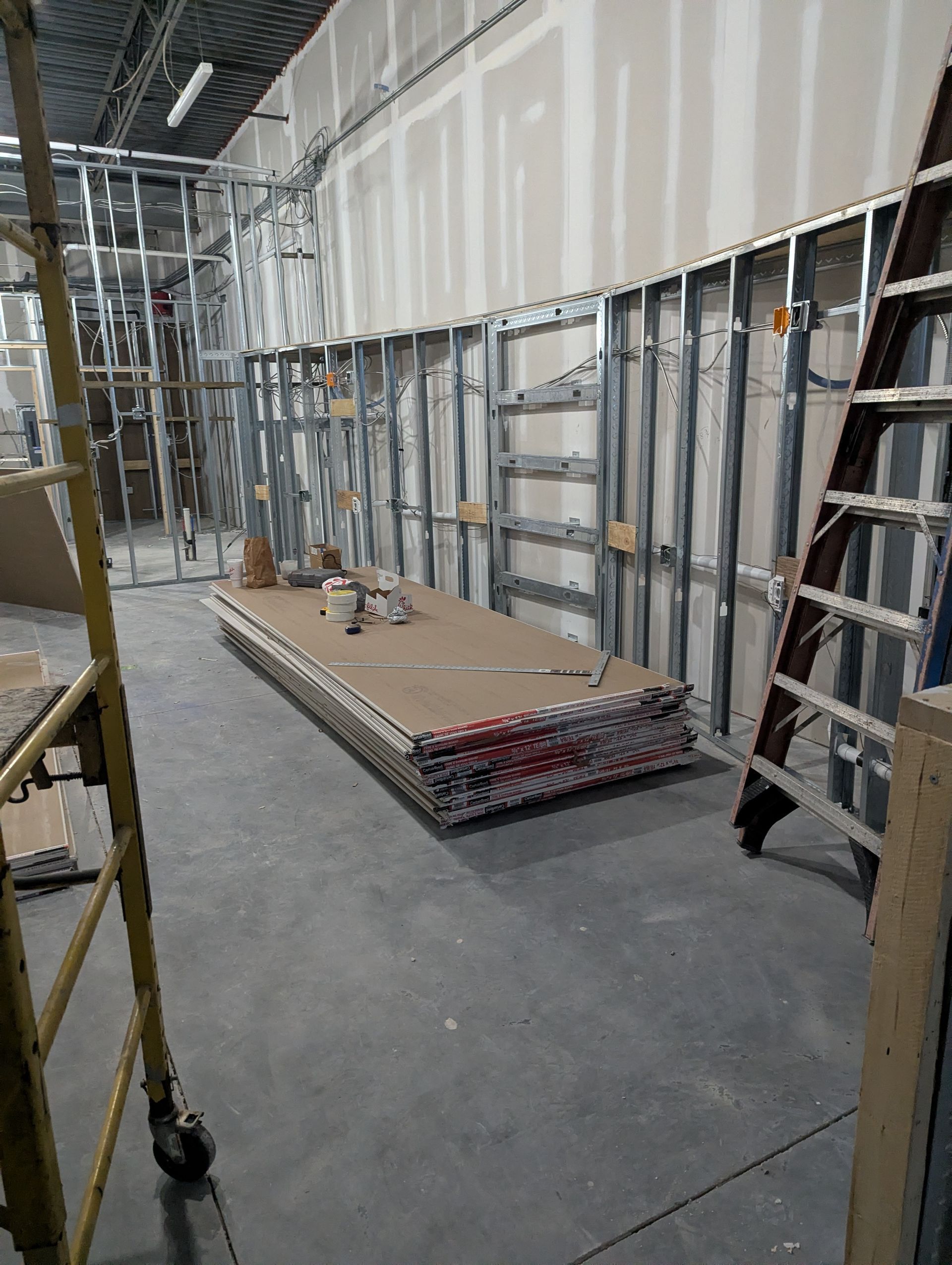 Construction interior showing a metal-framed wall, a stack of drywall sheets, and a ladder on a concrete floor.
