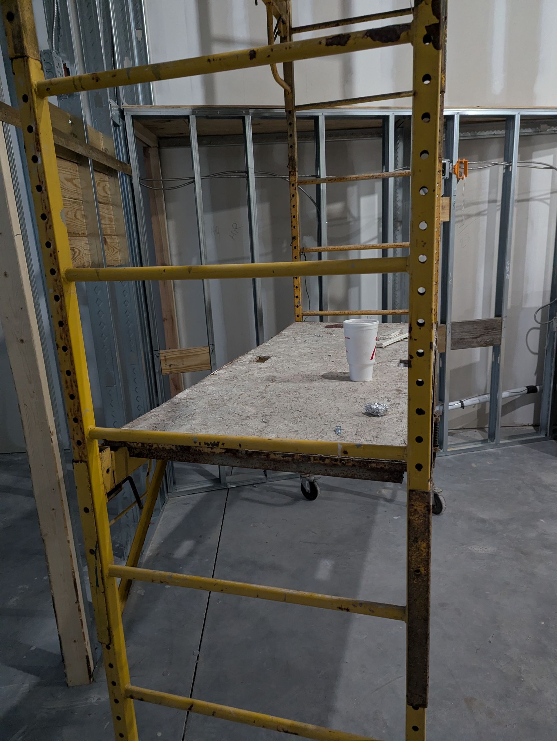 A yellow metal scaffolding unit stands in a room with unfinished metal wall studs, with a drink cup resting on its platform.