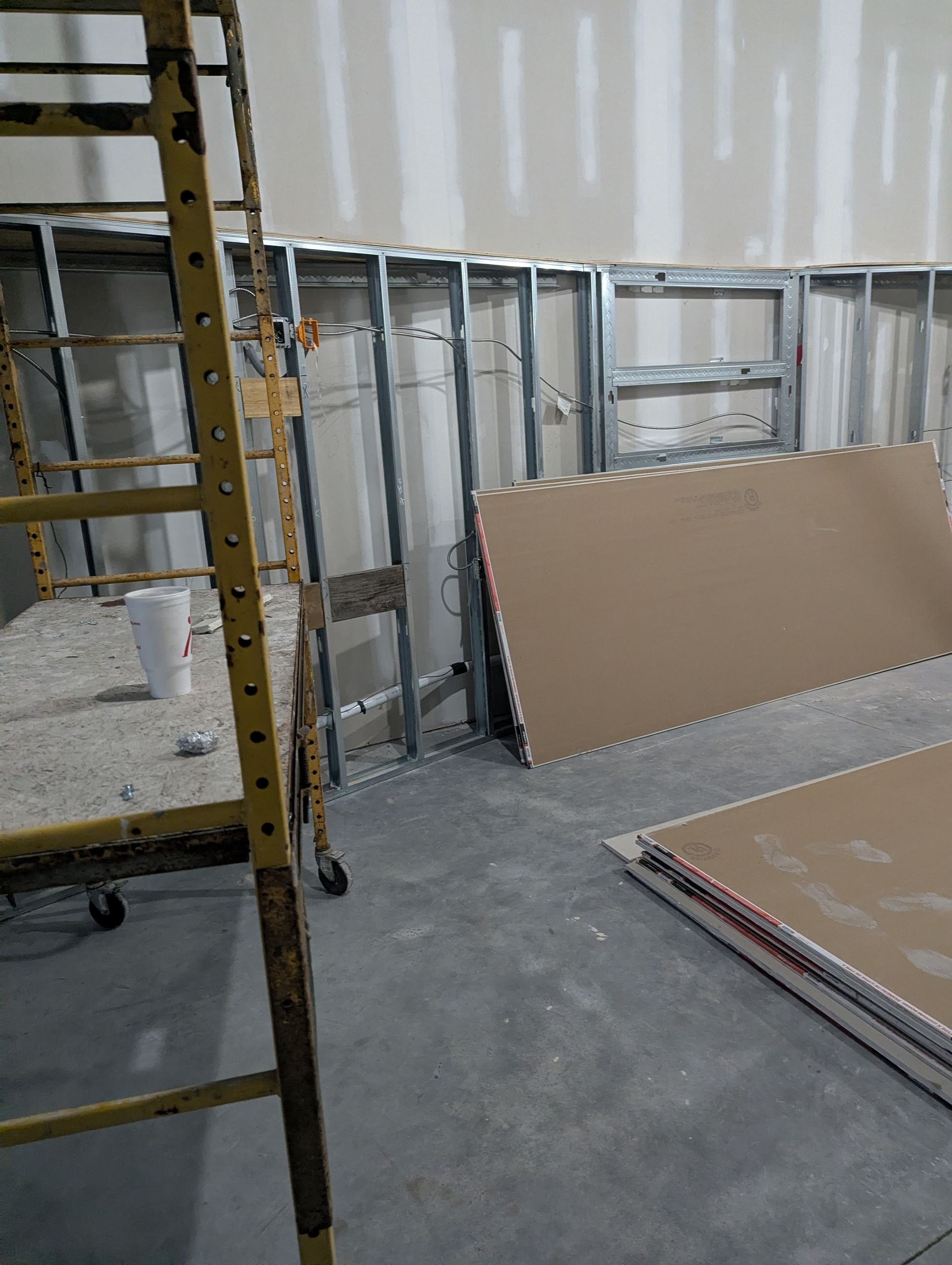 Construction scene showing metal wall studs, drywall panels, and scaffolding on a concrete floor.