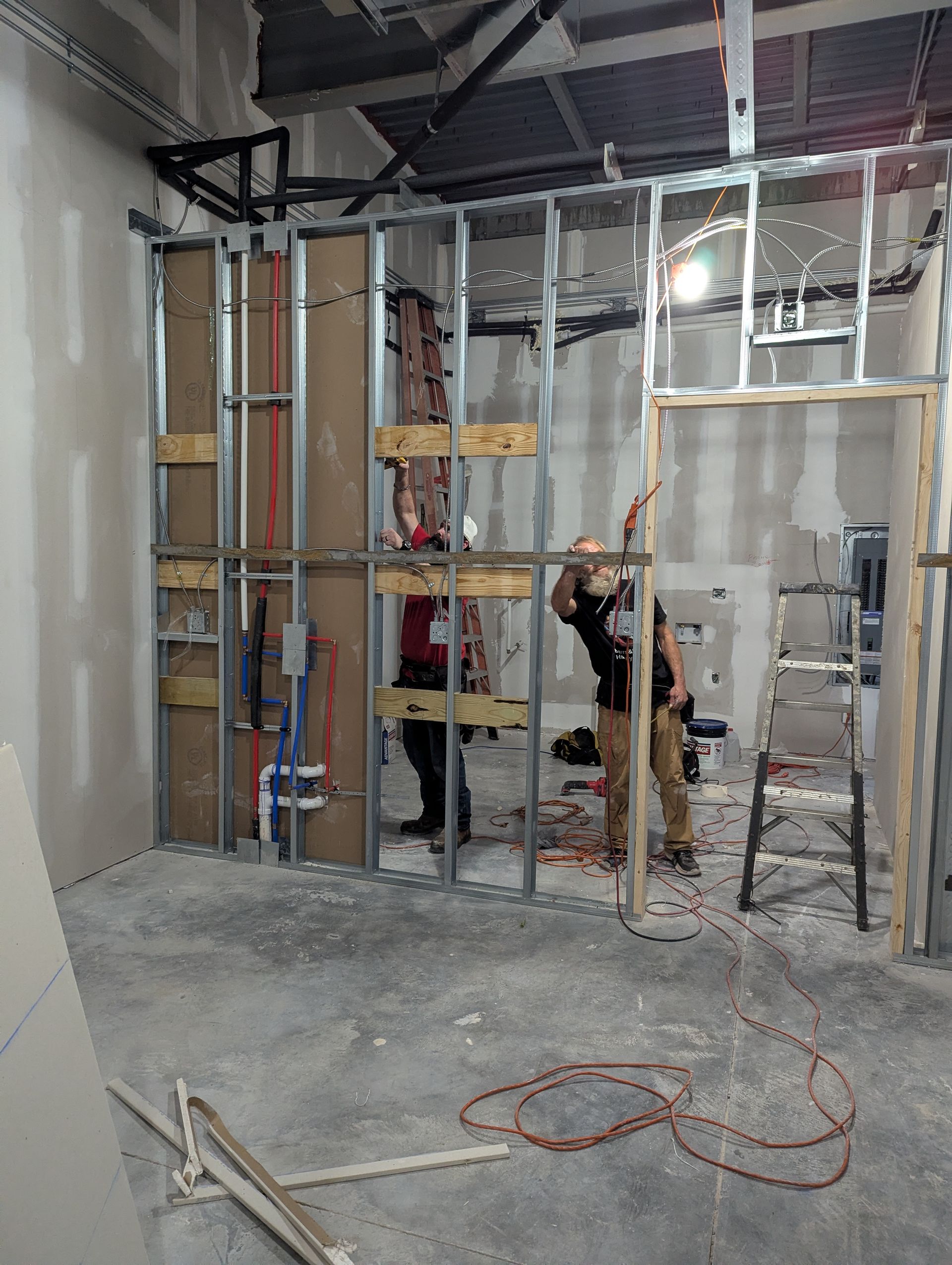Construction workers in an interior framing space with exposed metal studs, pipes, and a ladder.