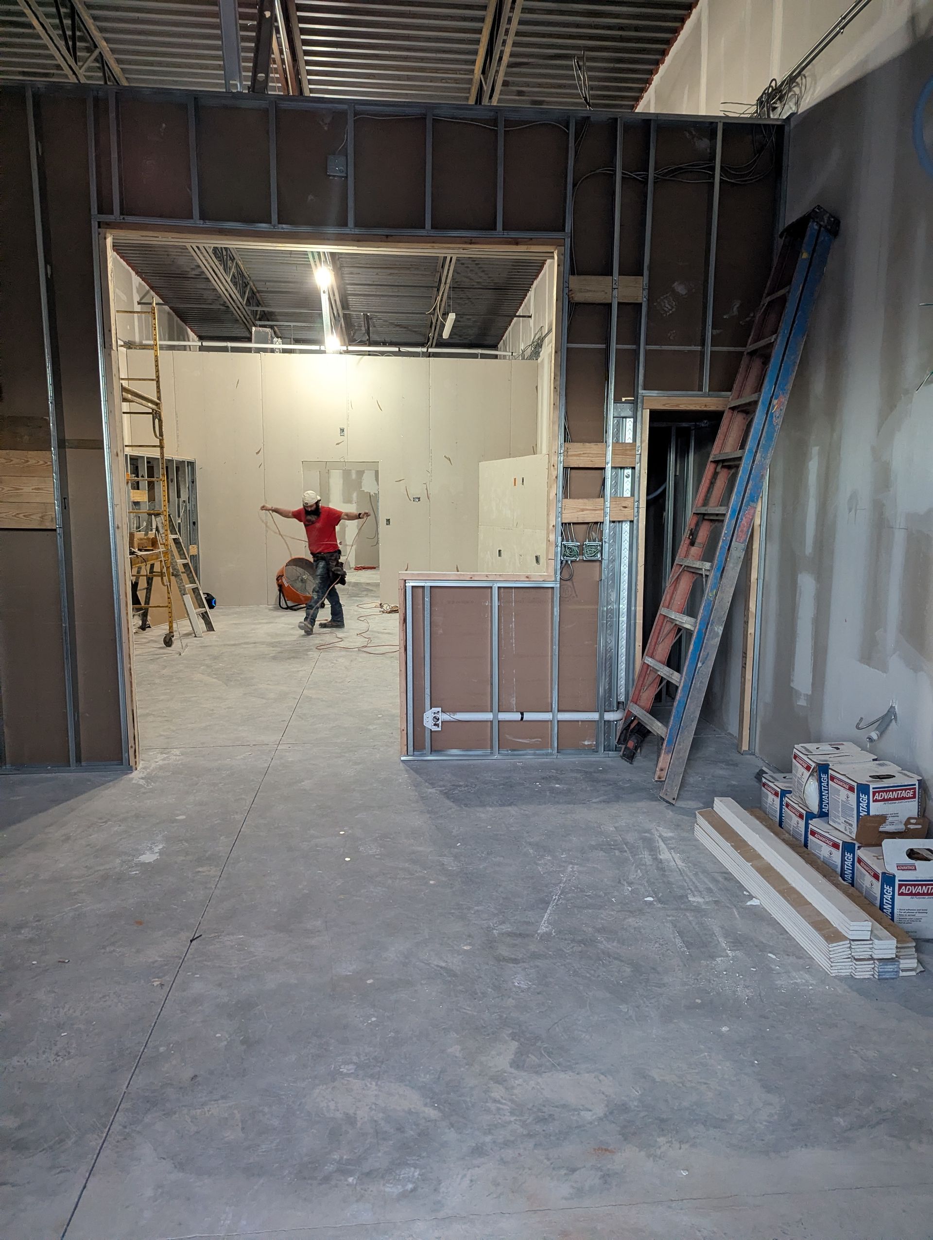 A worker in a red shirt stands in a construction site frame, with a blue ladder leaning against the wall nearby.