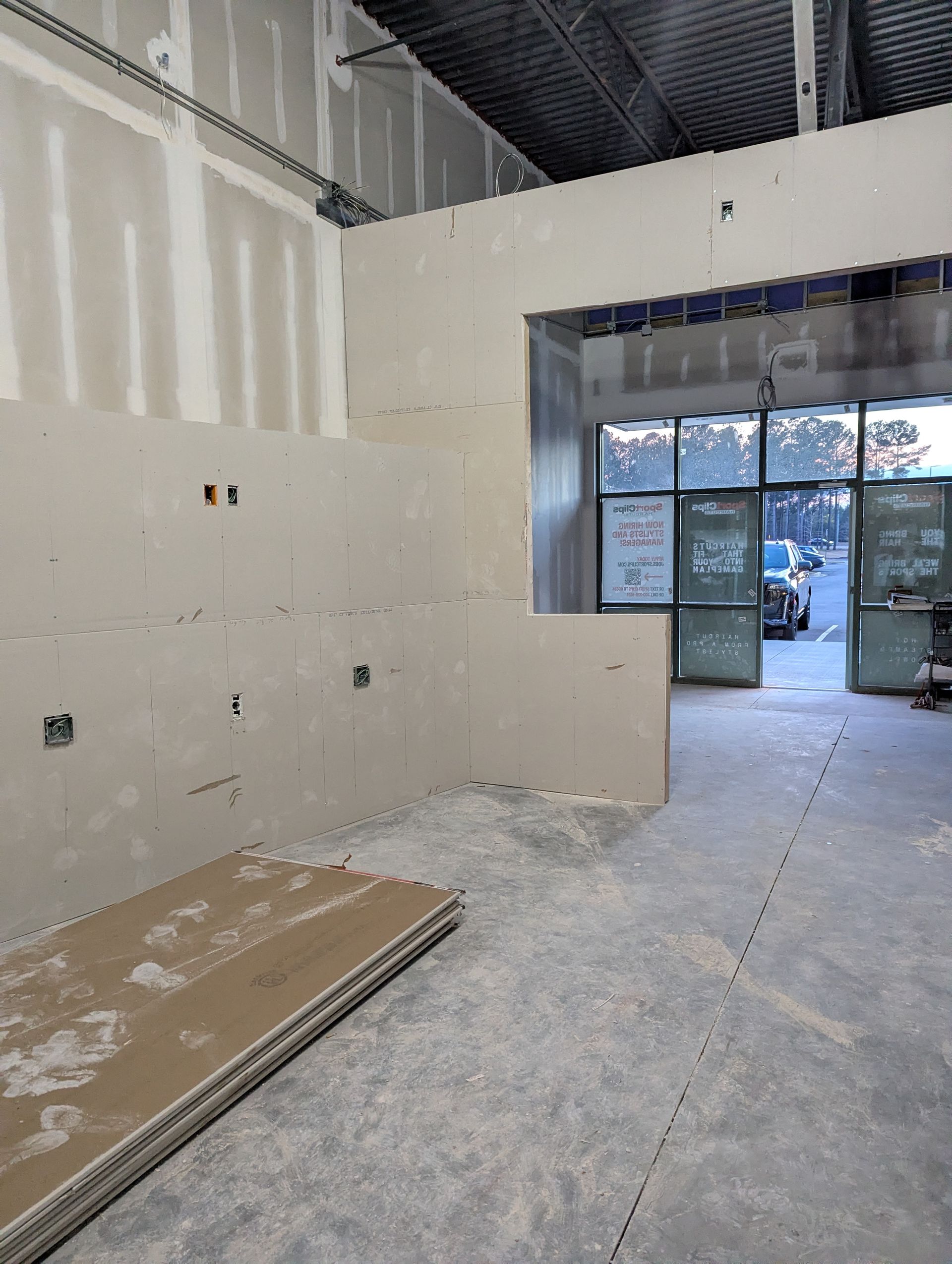 An interior construction site featuring unfinished drywall walls, exposed electrical boxes, and an entrance with glass doors.
