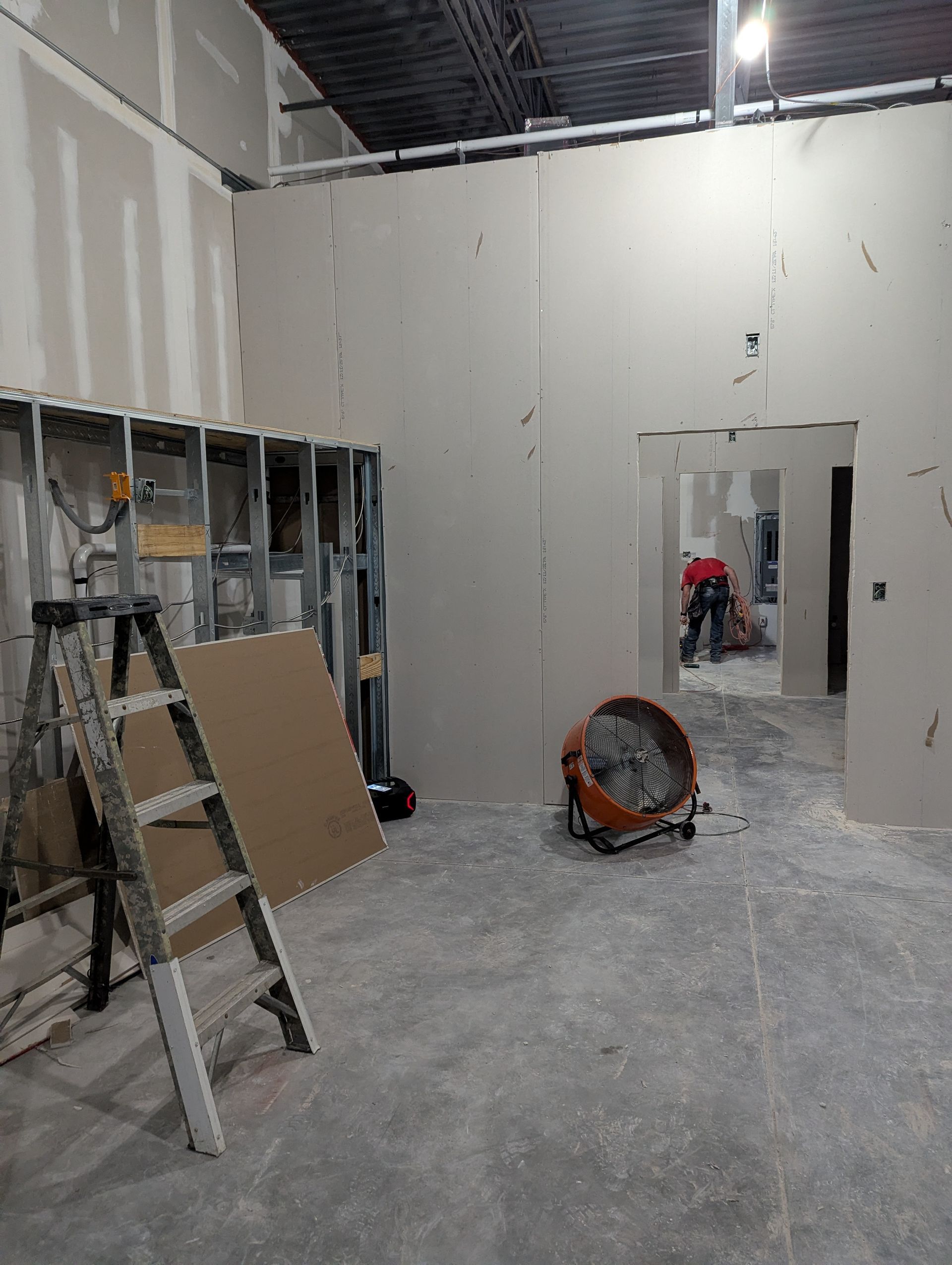 Interior construction site featuring drywall panels, a metal stud wall with a ladder, and a worker in an adjacent room.
