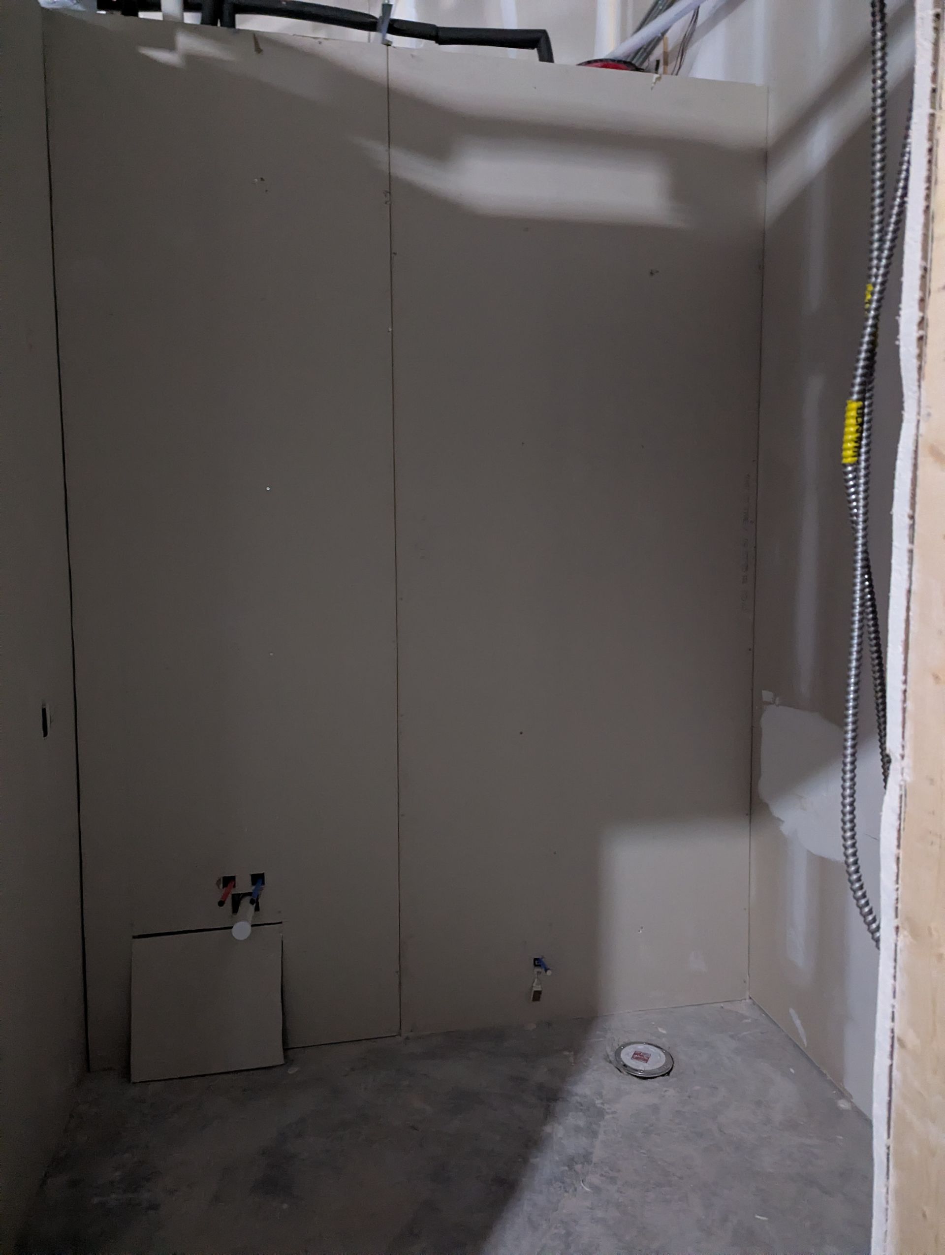 An unfinished interior space with drywall walls, exposed pipes overhead, and plumbing rough-ins on the floor and wall.