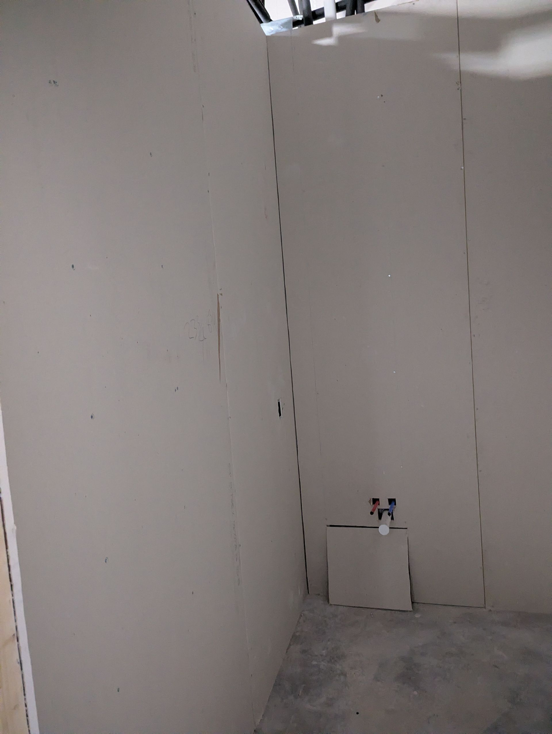 Unfinished drywall interior with a corner, a floor-level cutout, and visible plumbing pipes.