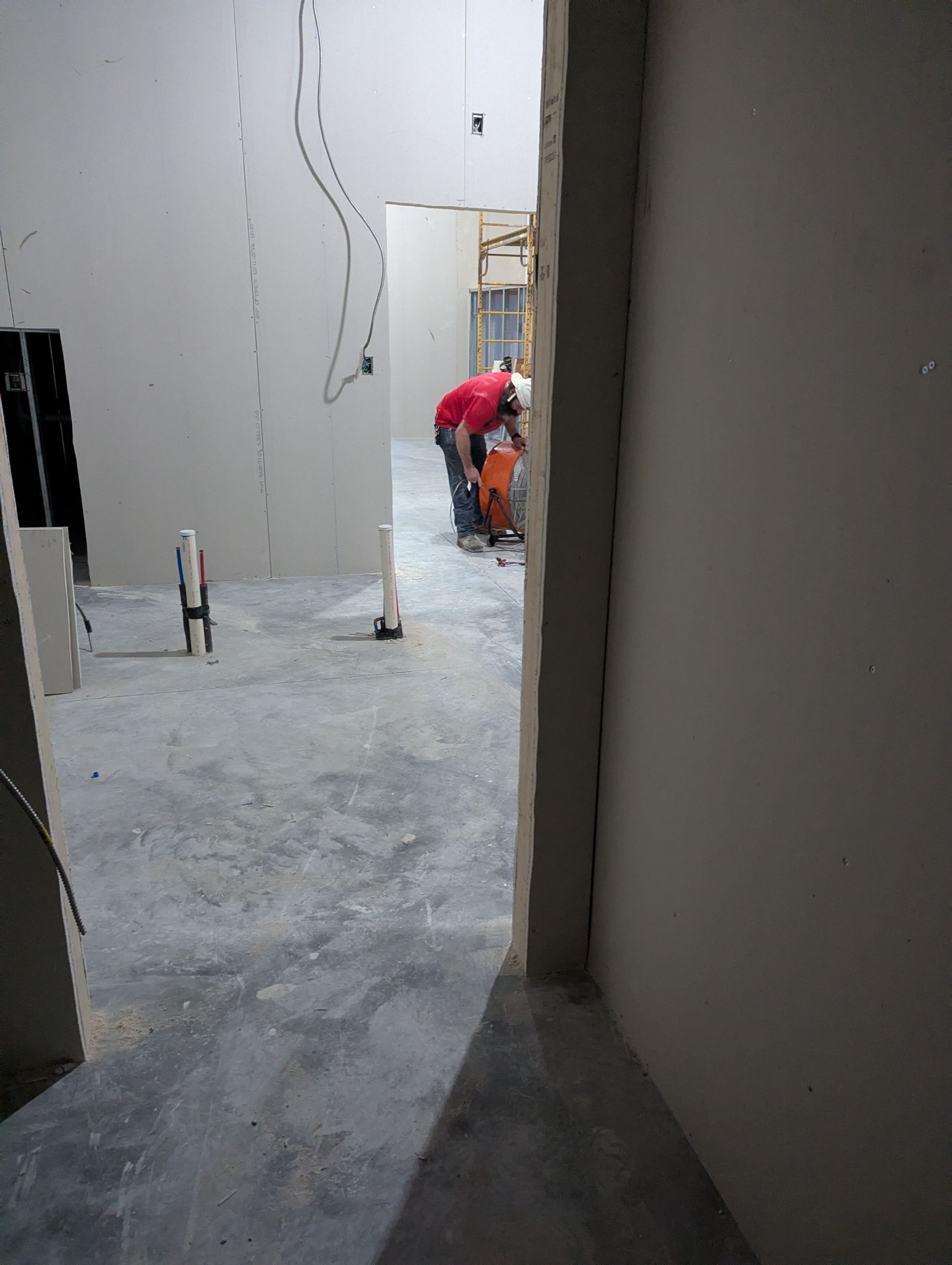 Construction worker in a red shirt cuts through concrete in an unfinished room with exposed pipes and drywall.