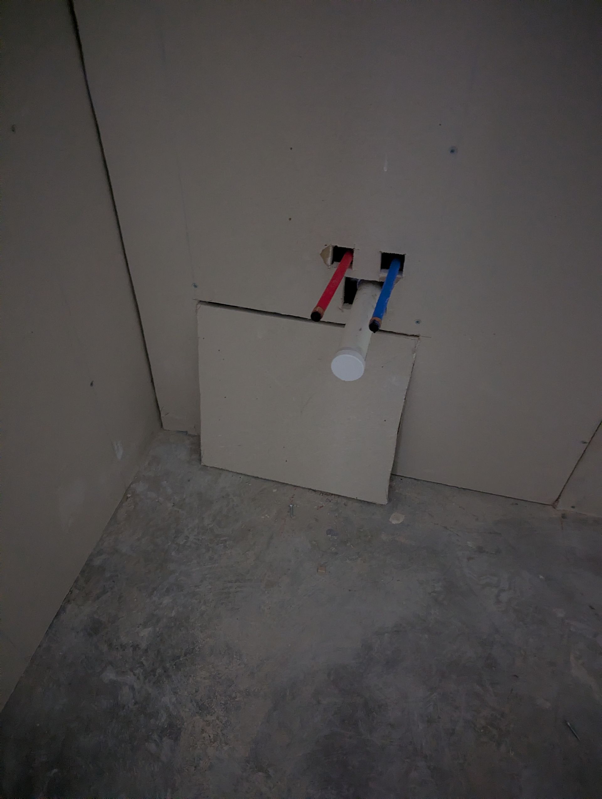 Construction site interior showing exposed red and blue water pipes extending from a patch in a drywall wall.