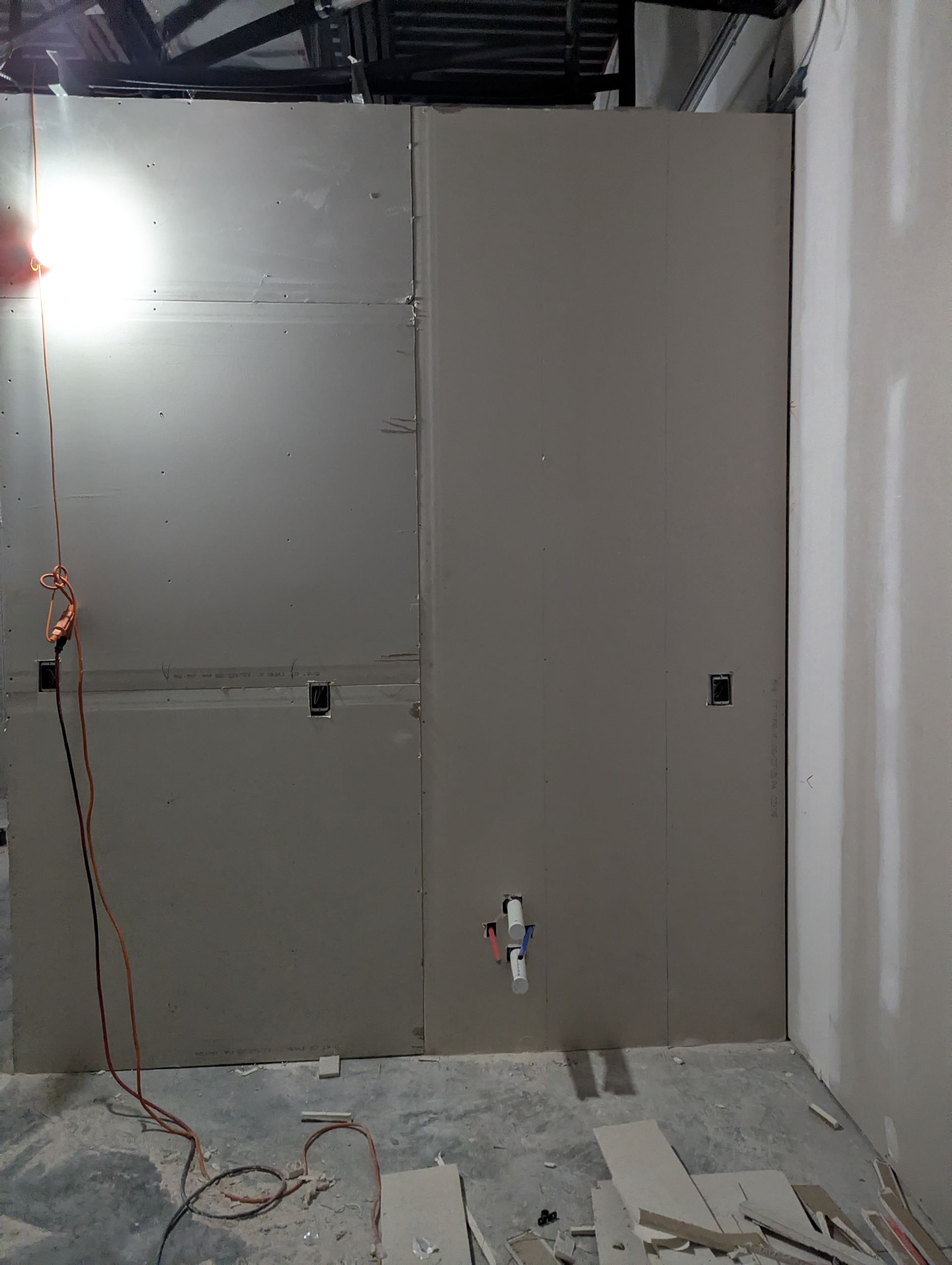 A partially installed drywall wall in an unfinished room with electrical outlets and a pipe protruding from the surface.