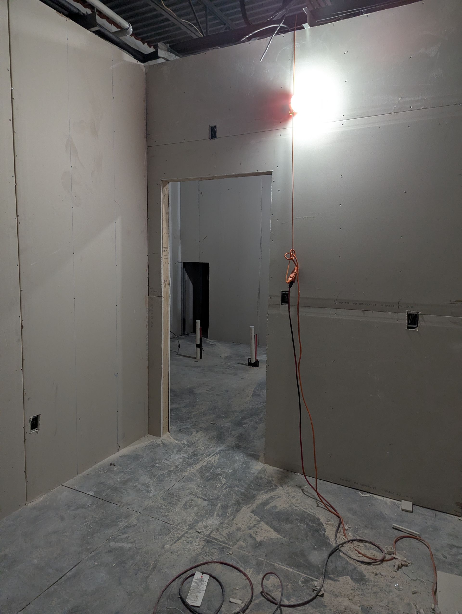 An interior construction site showing gray drywall walls, an open doorway, exposed ceilings, and electrical wiring.