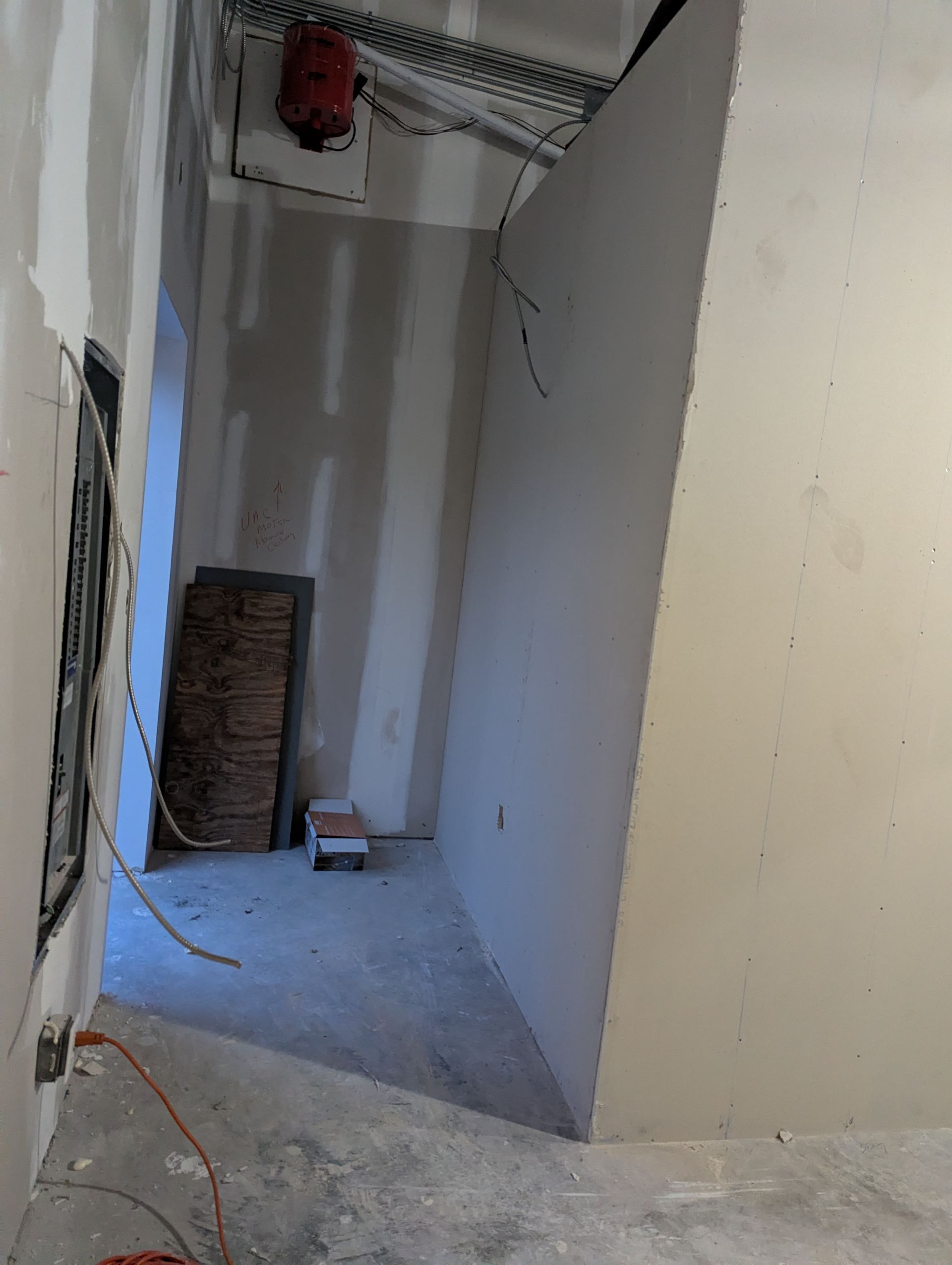 A view of an unfinished room with drywall, exposed wiring, a ceiling-mounted red ventilation unit, and construction debris.