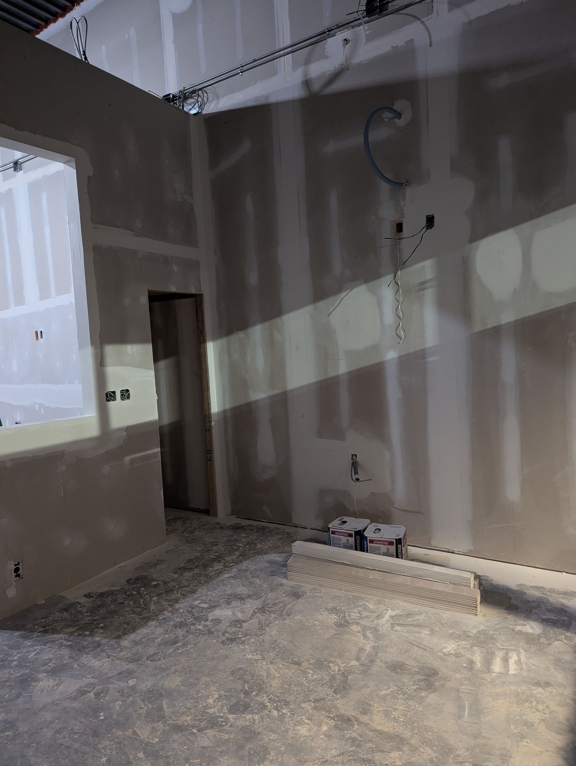 An unfinished room interior with drywall panels, exposed wires, and a doorway, viewed under construction lighting.
