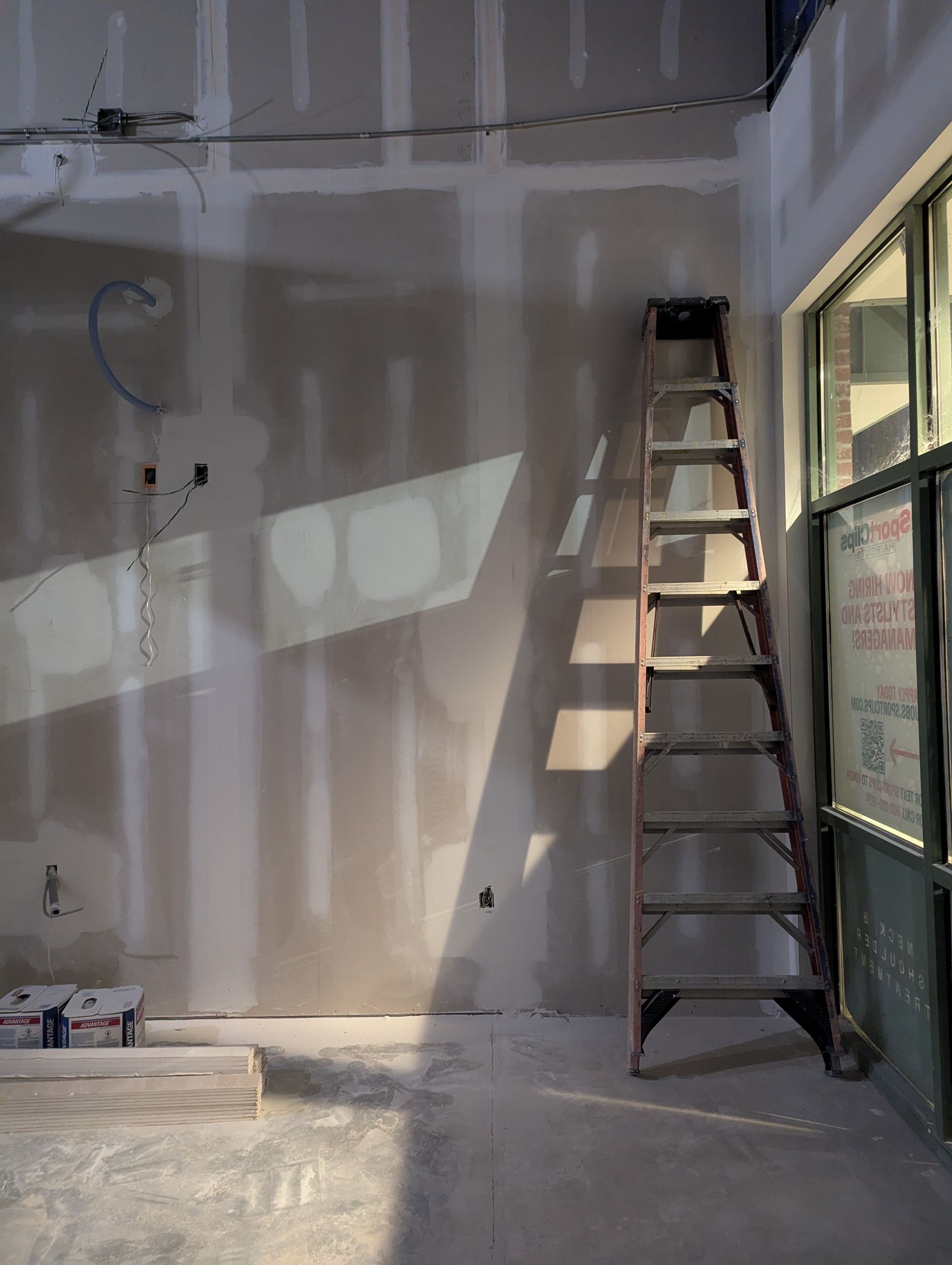 An interior view of a room under construction with unfinished drywall, an A-frame ladder, and bright natural sunlight.