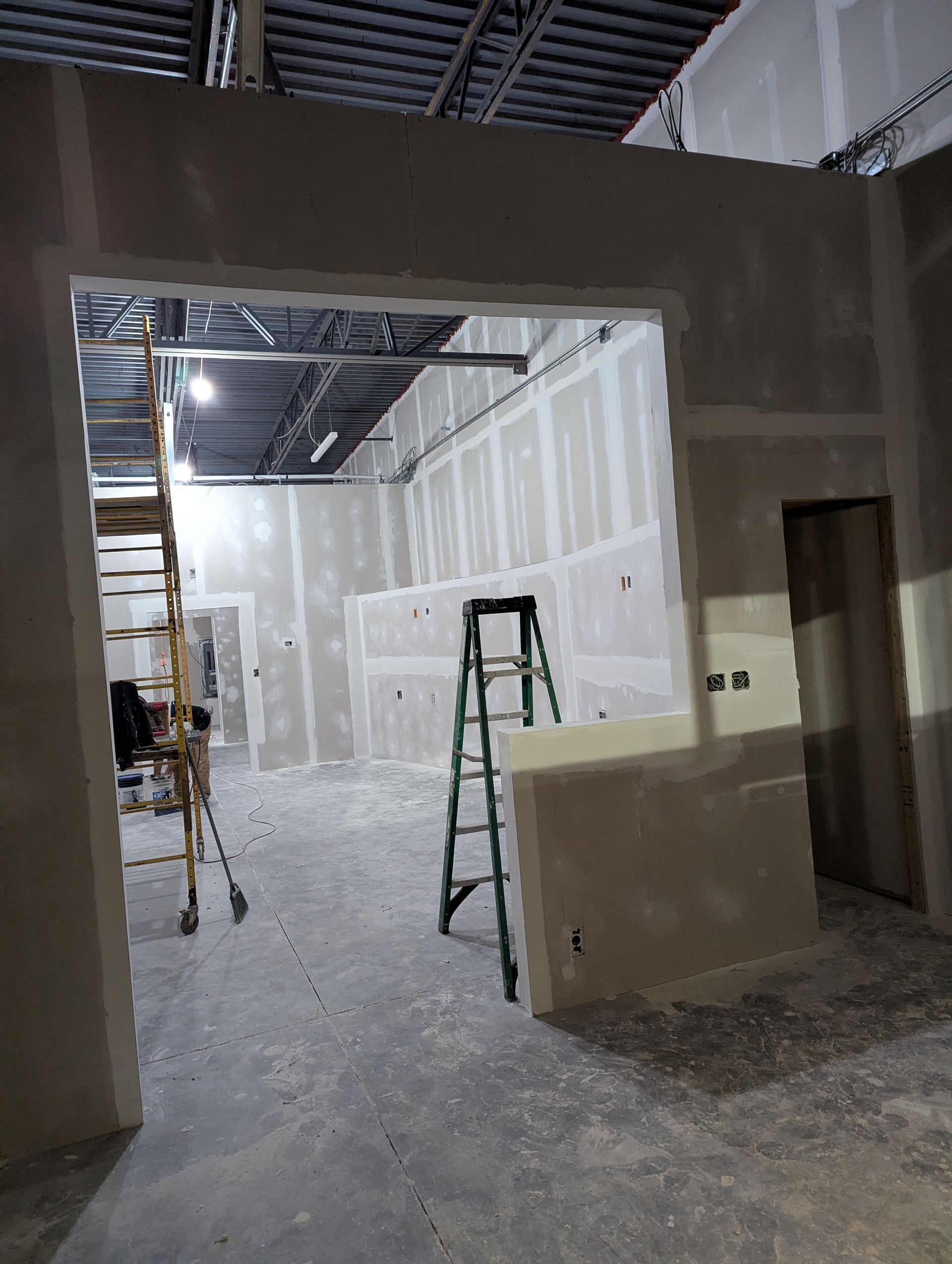 An unfinished commercial space with drywall, framing, a ladder, and rolling scaffolding, indicating ongoing construction.