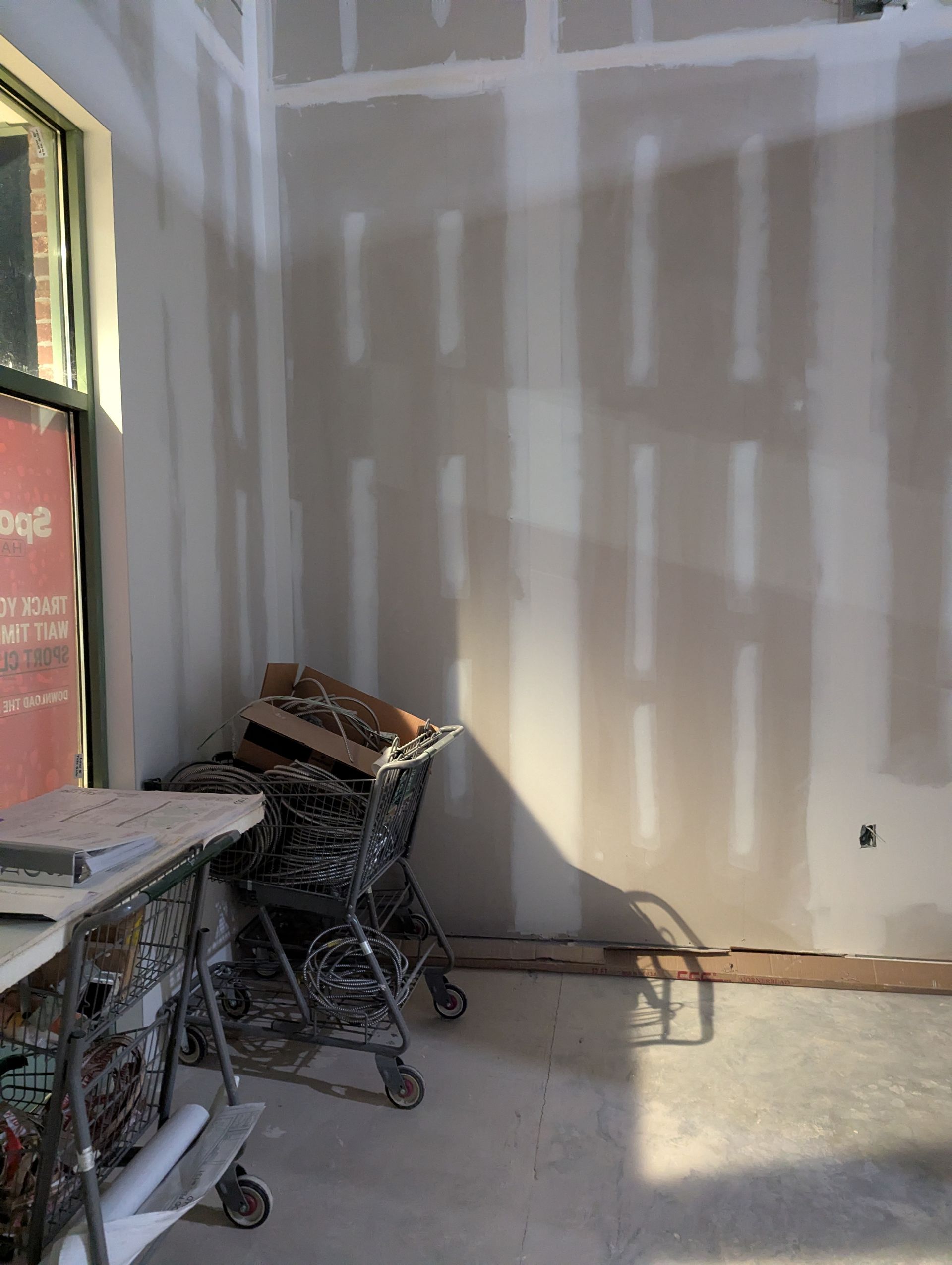 An interior room under construction with unfinished drywall, a window, and a utility cart filled with building supplies.