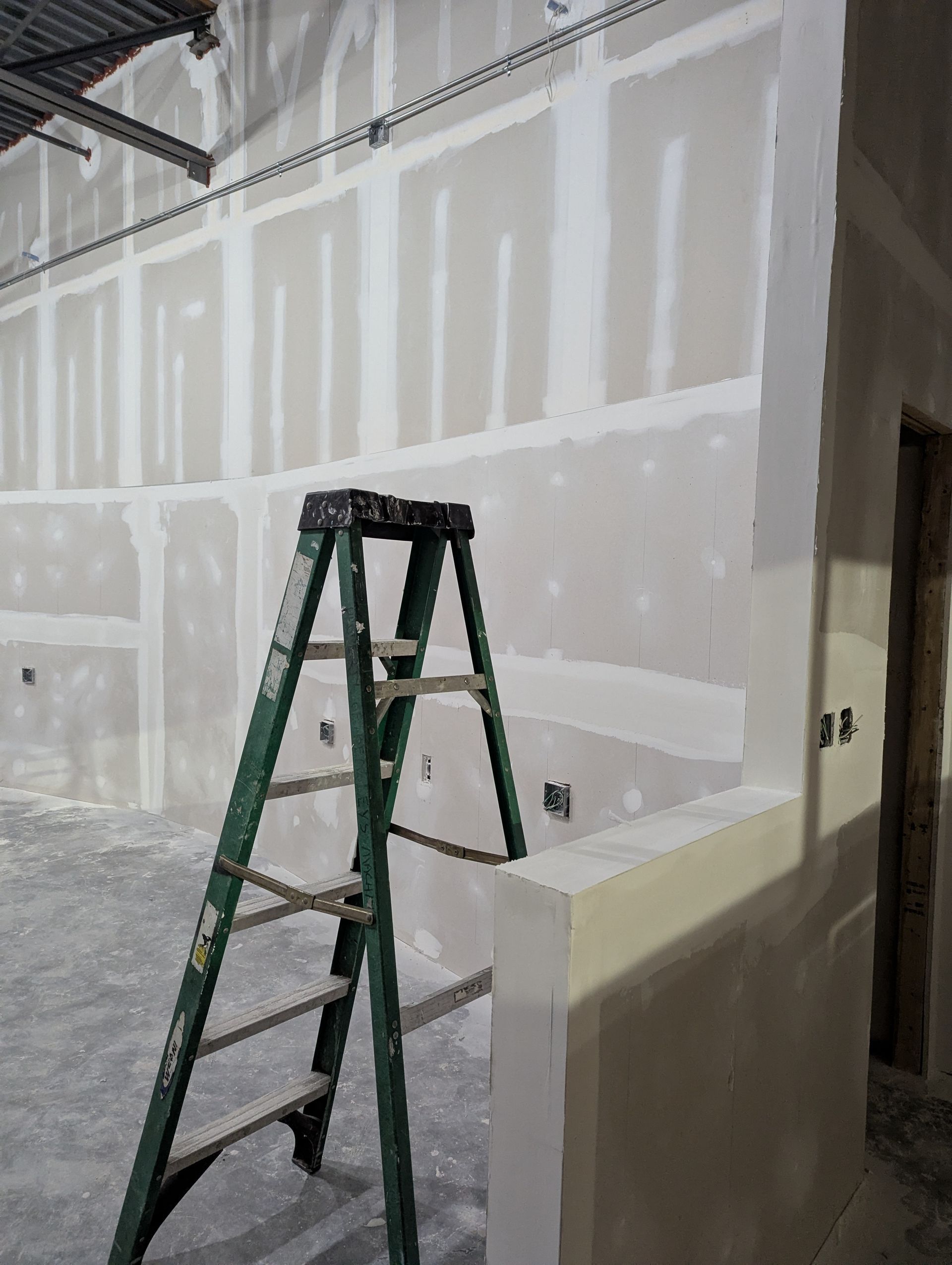 A green stepladder stands in a room with unfinished drywall walls featuring taped joints and spackled screw holes.
