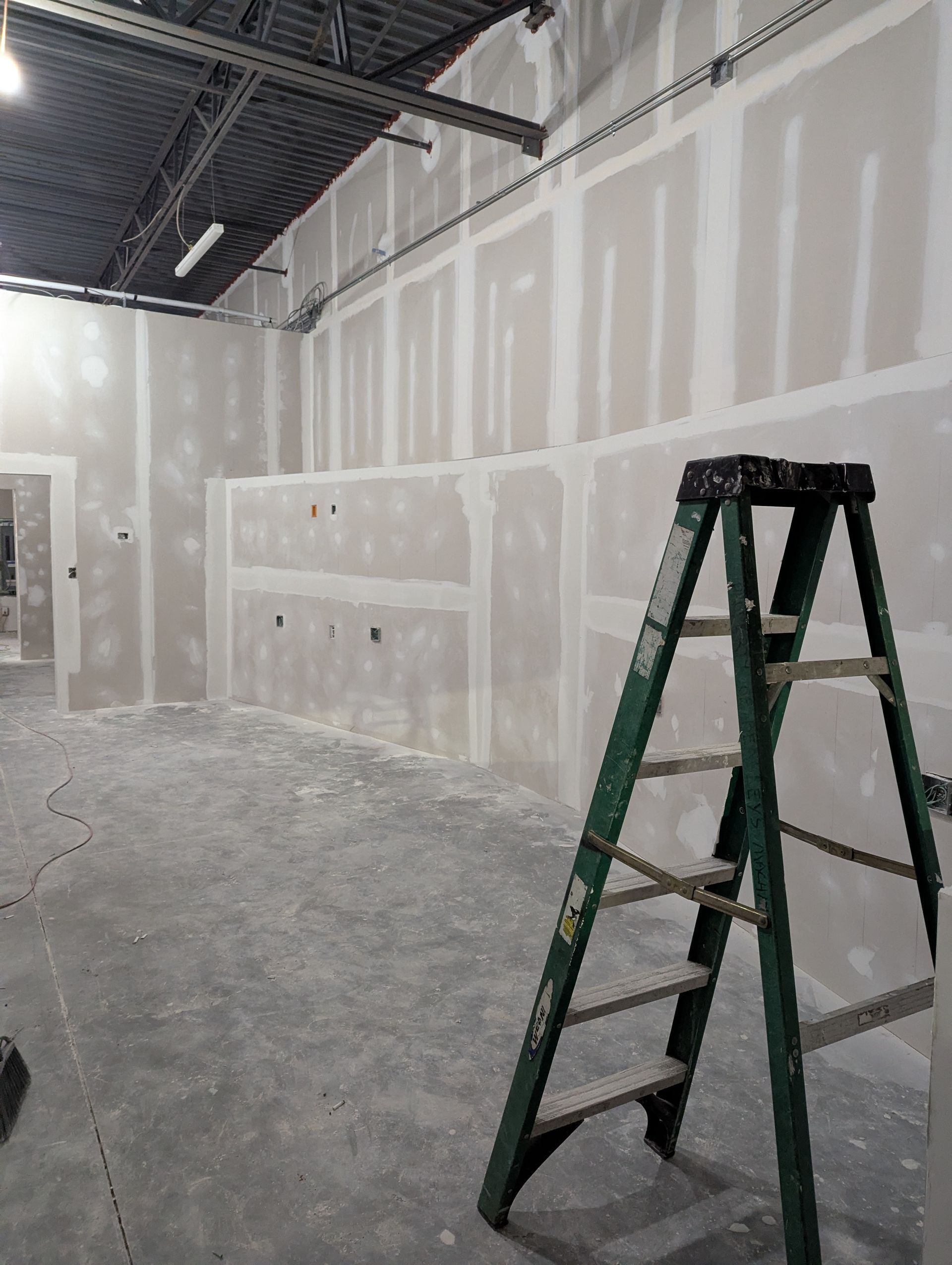 An interior construction site showing freshly installed drywall with taped joints and a green stepladder in the foreground.