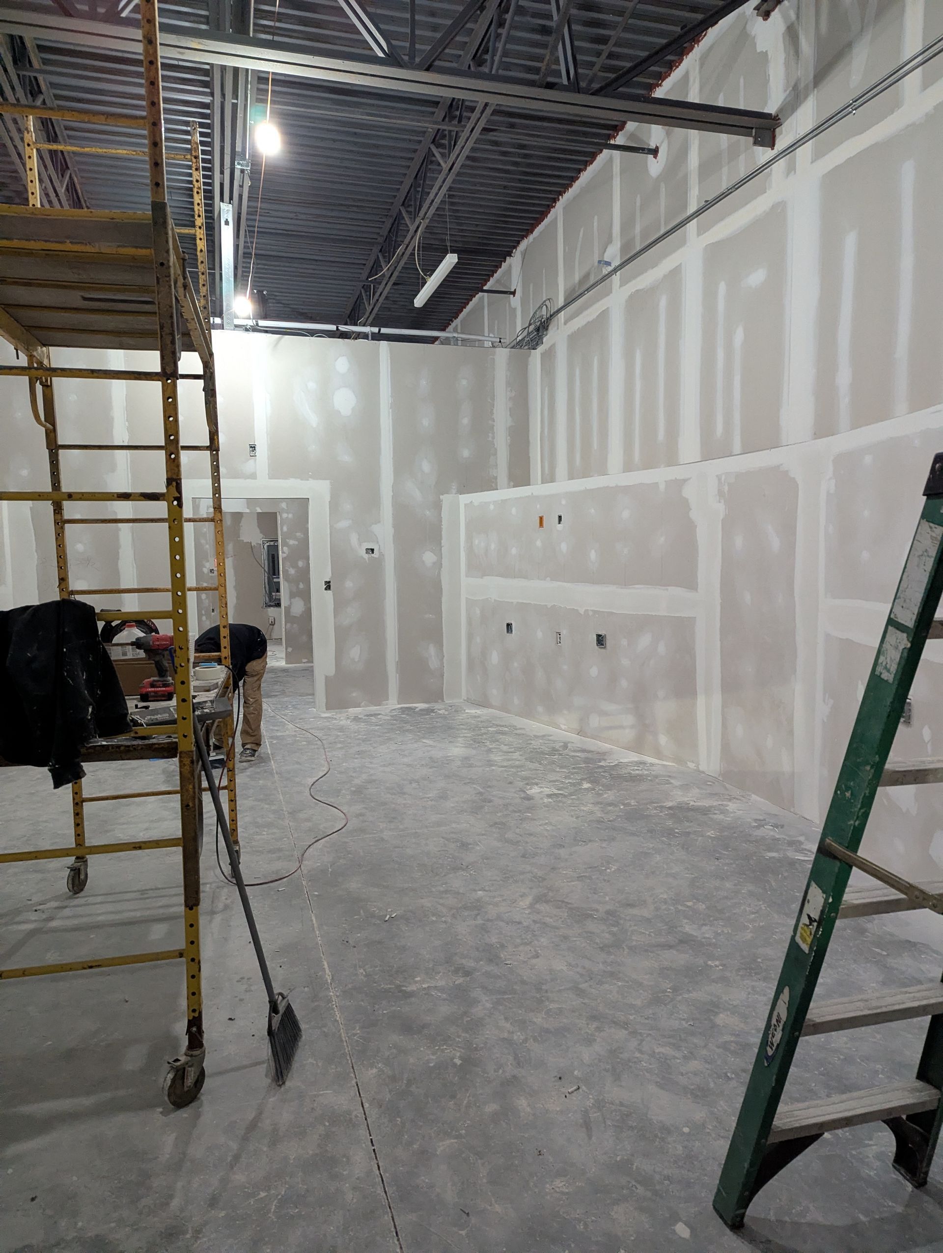 A construction worker inside a room with unfinished drywall walls, a rolling scaffold, and a stepladder.