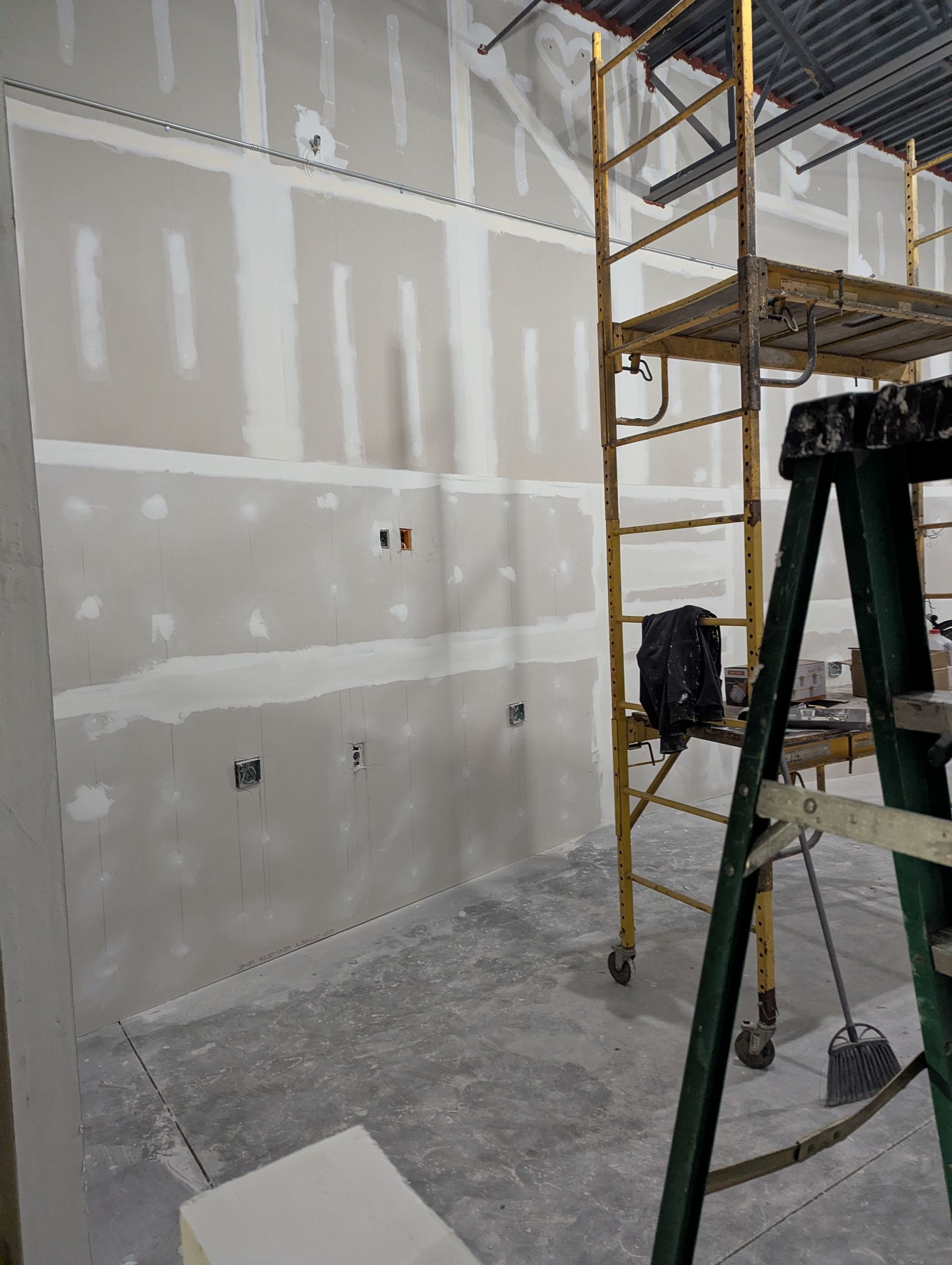 An interior view of a room under construction featuring drywall panels with taped seams and metal scaffolding.