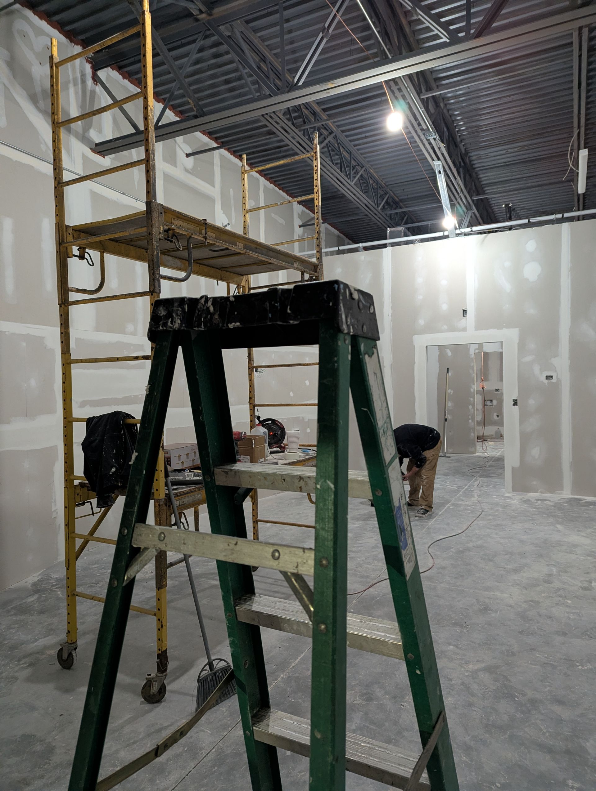 A dark green A-frame ladder sits in the foreground of a construction site with drywall walls and a tall yellow scaffold.
