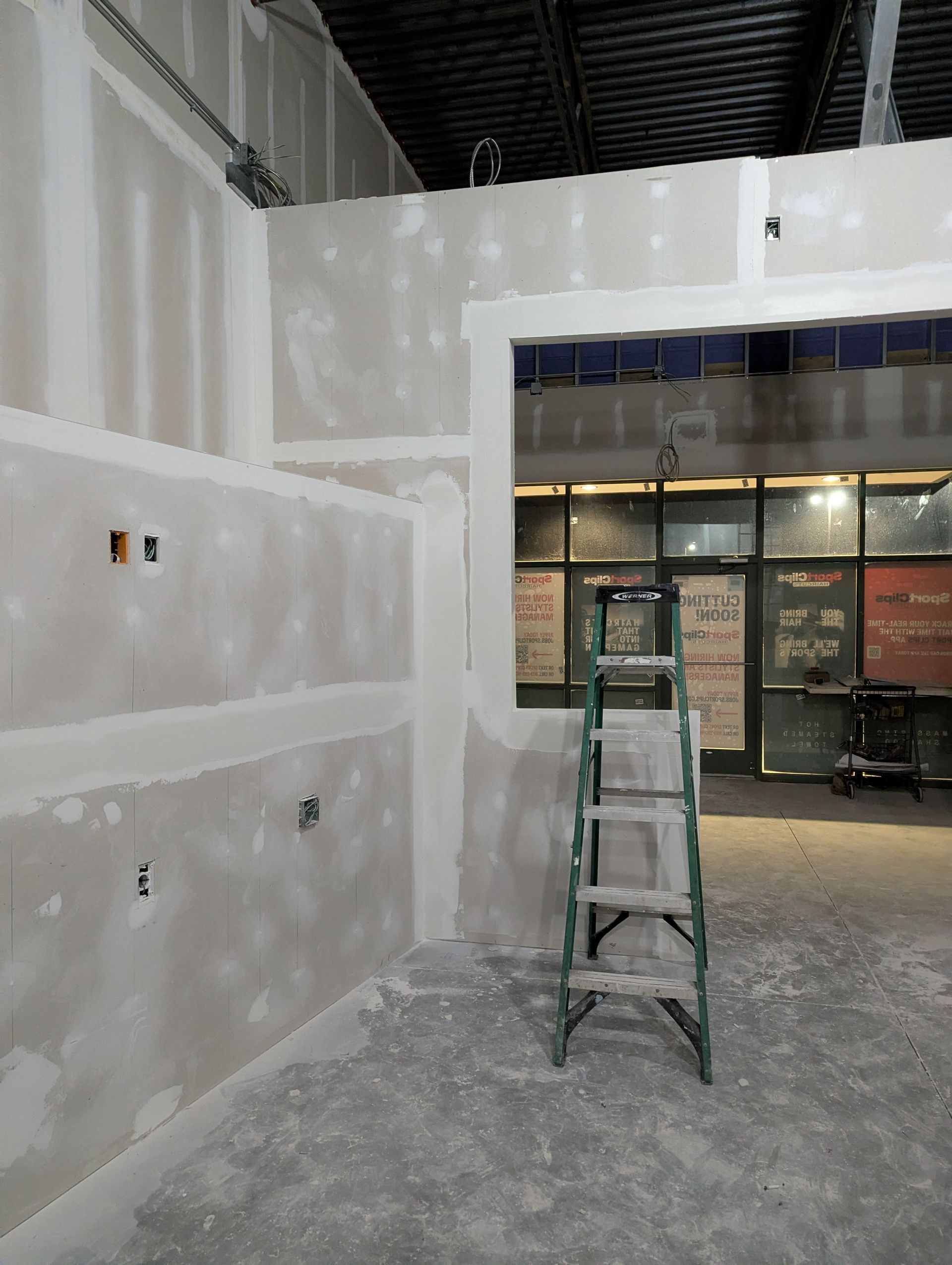 A ladder stands inside an unfinished room with newly installed, taped drywall, facing a storefront with glass windows.