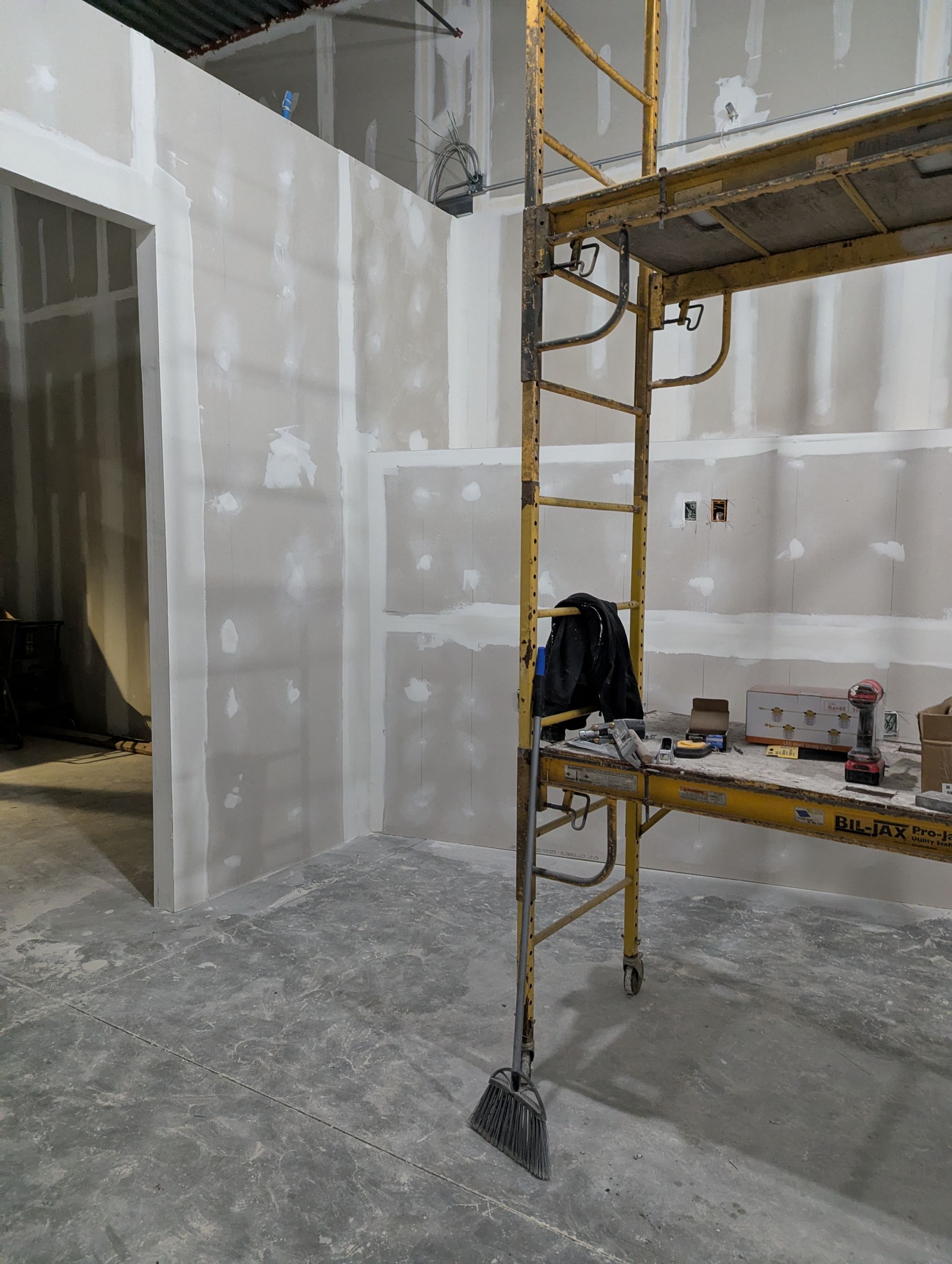 An interior construction site features unfinished drywall walls and a tall yellow metal scaffolding unit.
