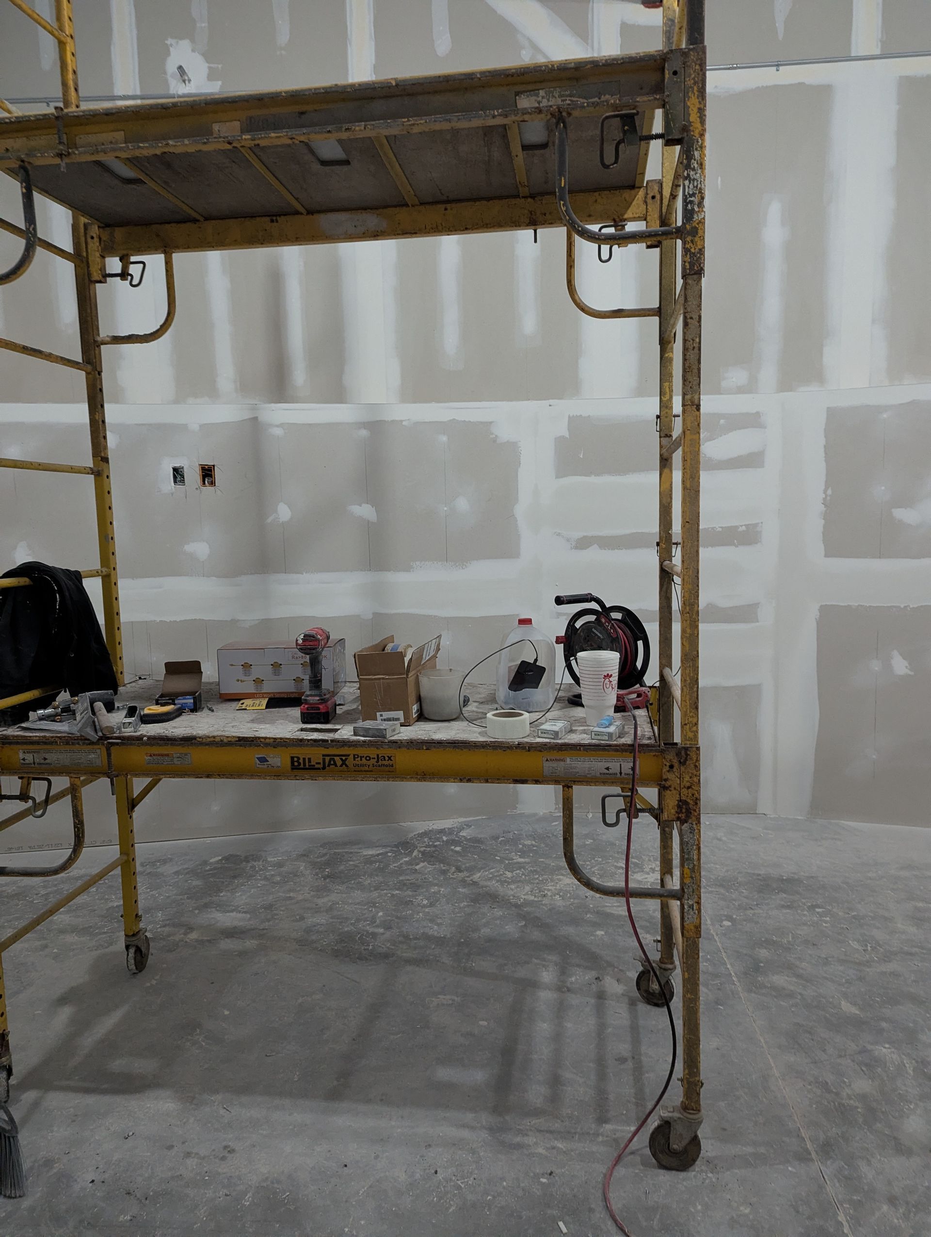 A yellow rolling construction scaffold sits in a room with unfinished, taped drywall walls and a concrete floor.