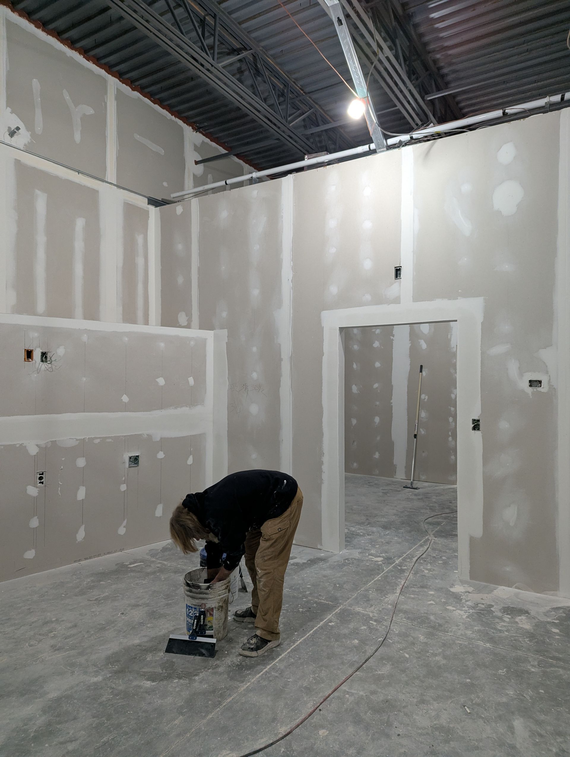 A person wearing a dark jacket and tan pants works on the floor of a room with unfinished drywall and a doorway.