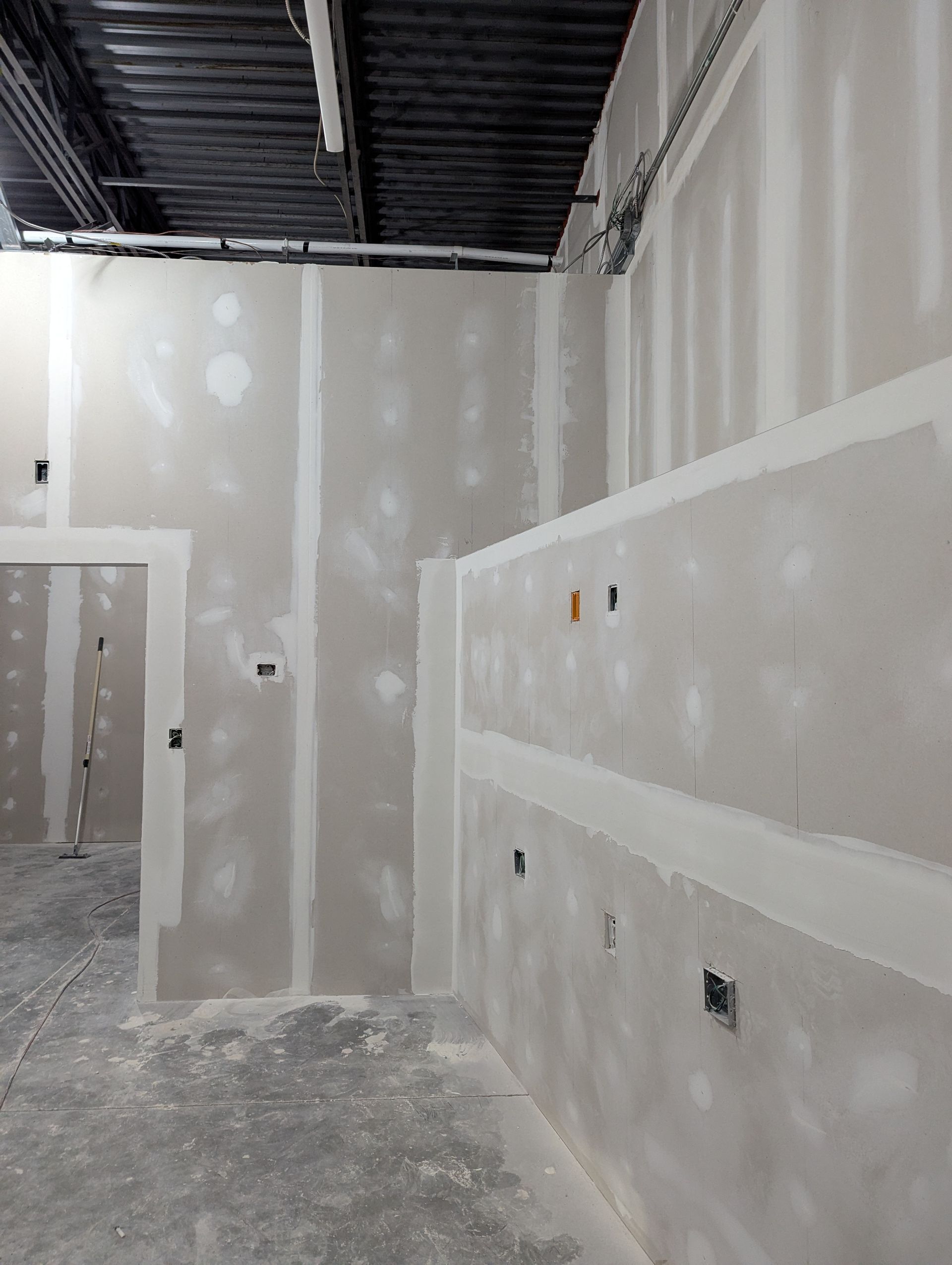 An interior construction site with unfinished, light-colored drywall walls and exposed black ceiling joists.