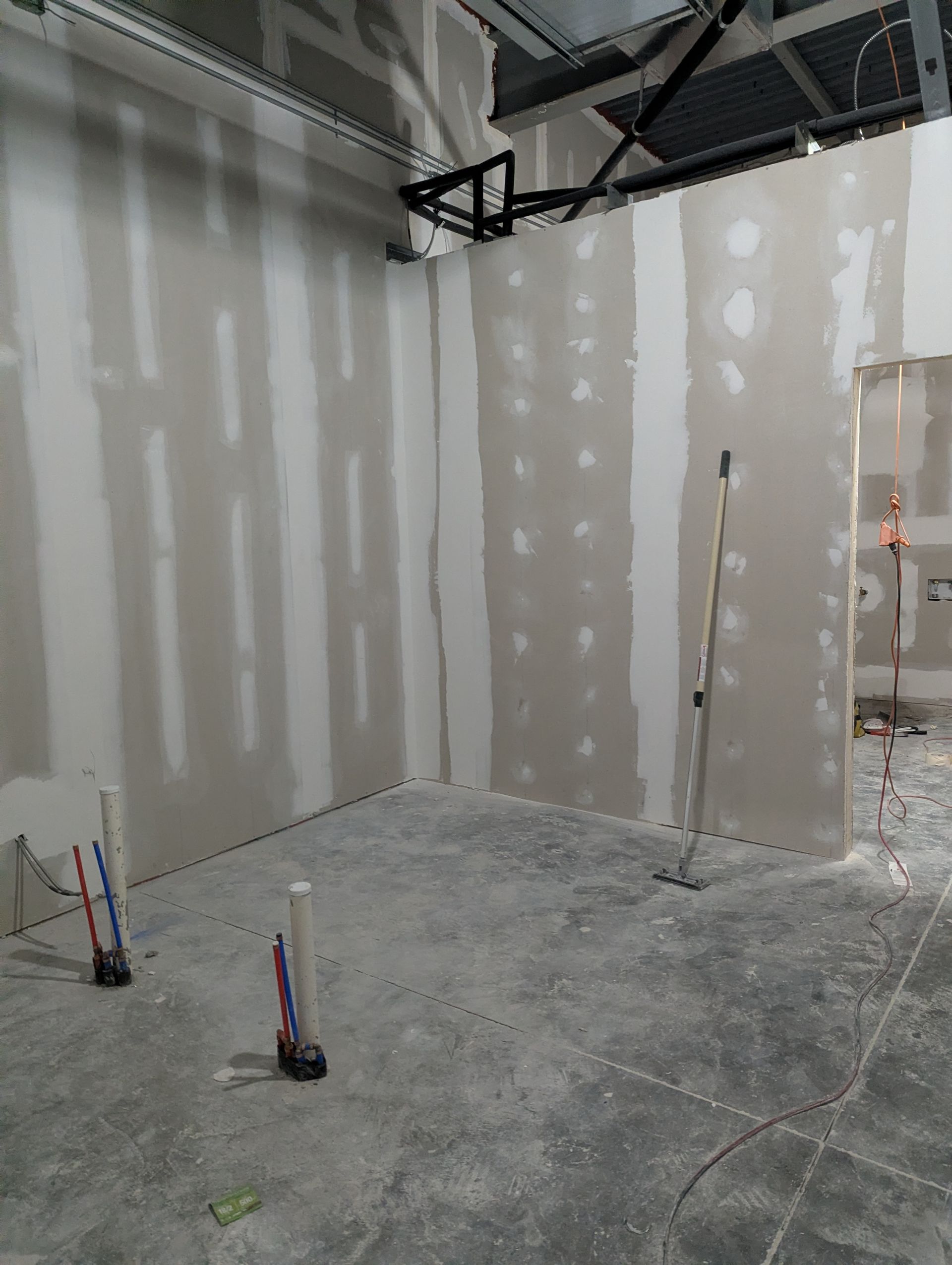 An unfinished interior room with drywall panels, exposed ceiling pipes, and utility pipes protruding from a concrete floor.