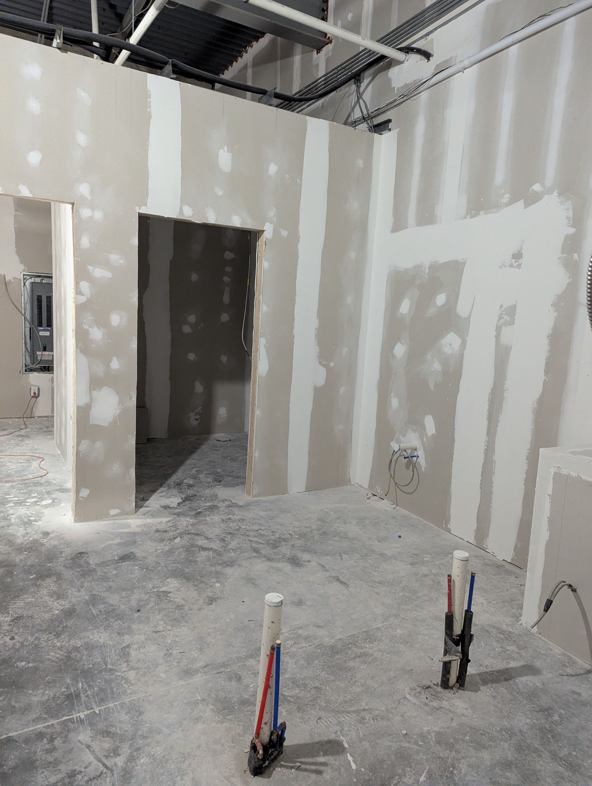 An interior room under construction with drywall, mudded seams, exposed ceiling pipes, and floor plumbing stub-outs.