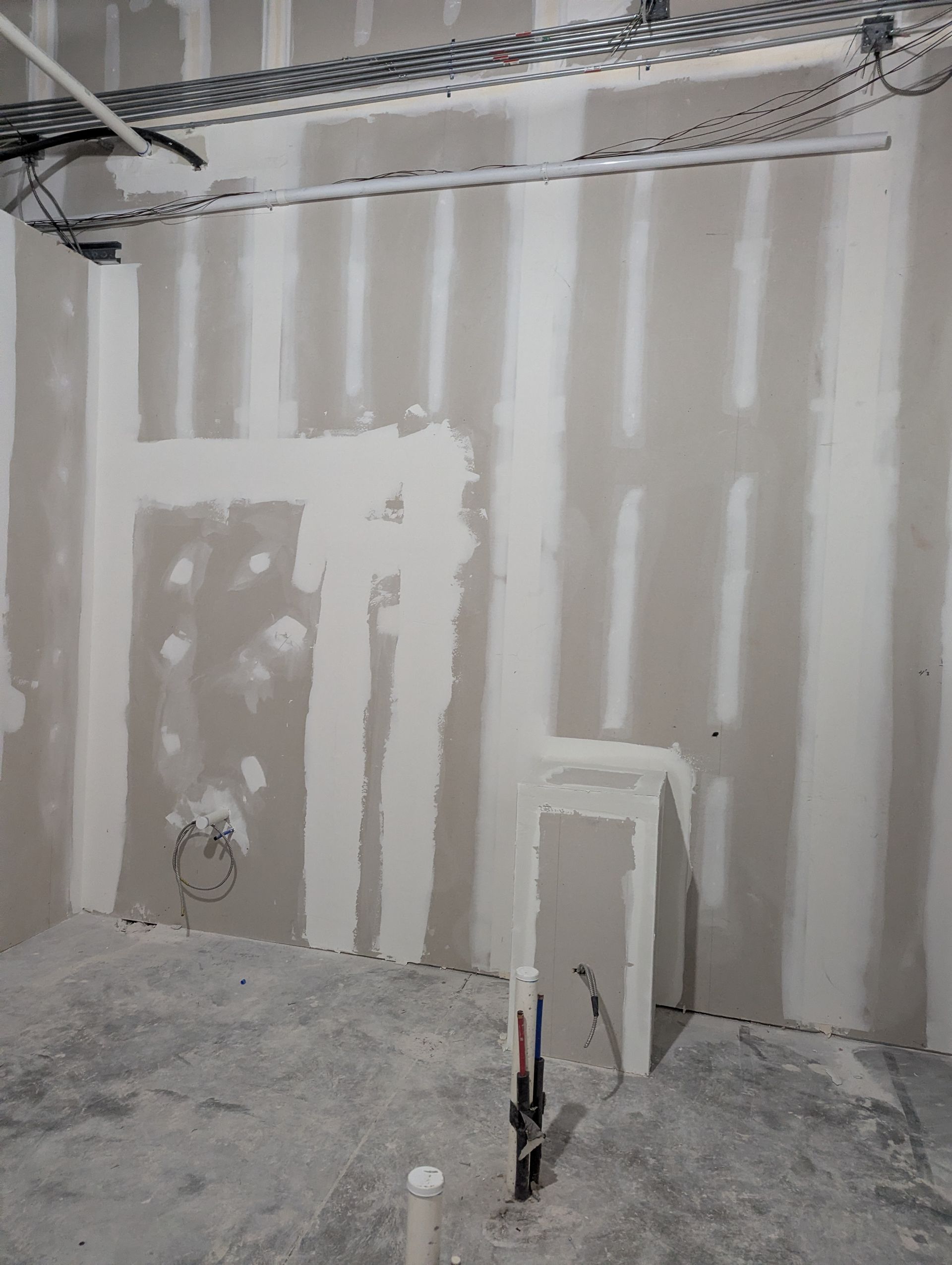 An unfinished room with drywall partitions, exposed ceiling wiring, and plumbing pipes protruding from the floor.