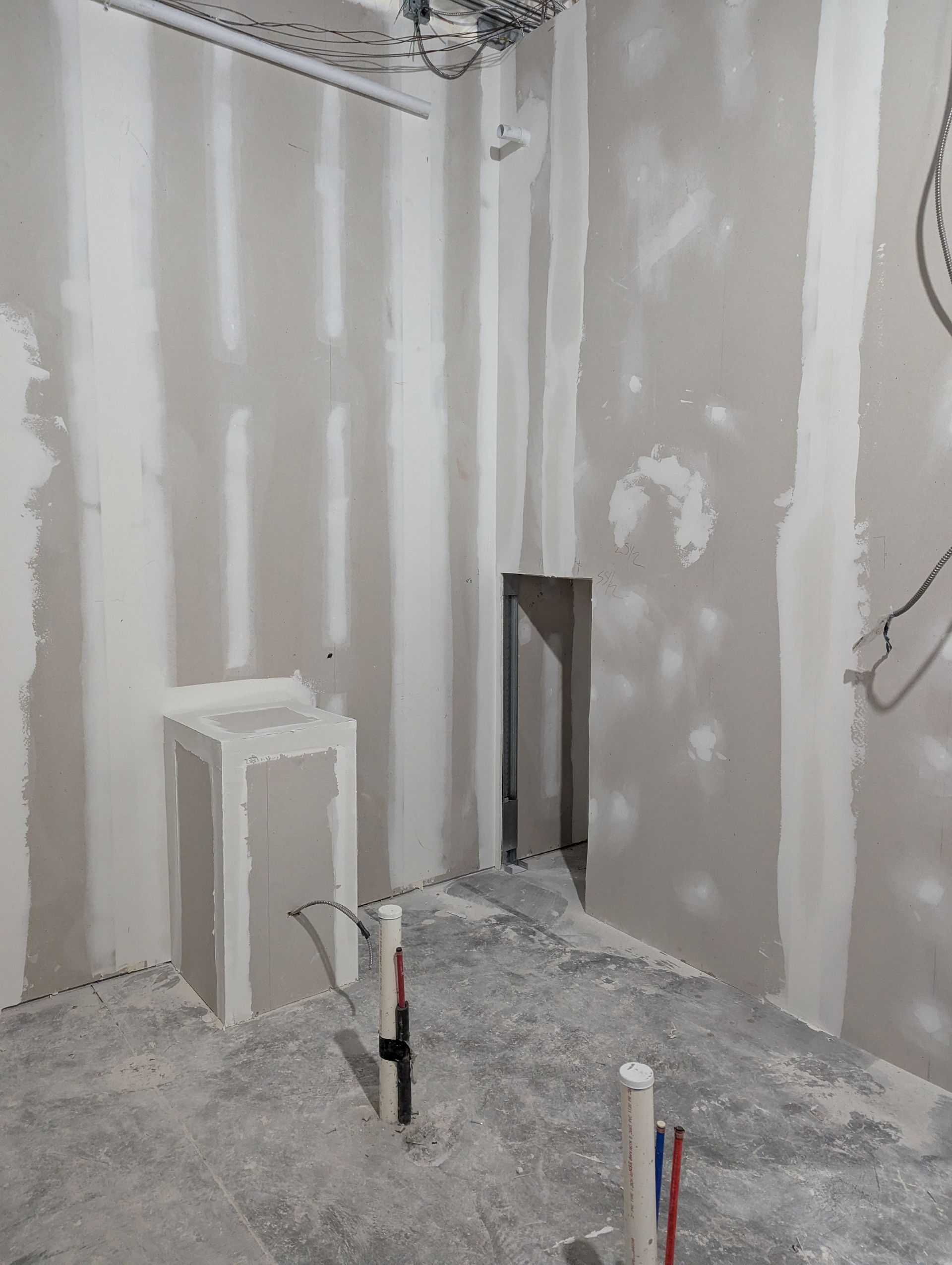An unfinished interior room with gray drywall, exposed plumbing pipes, and a small open doorway.