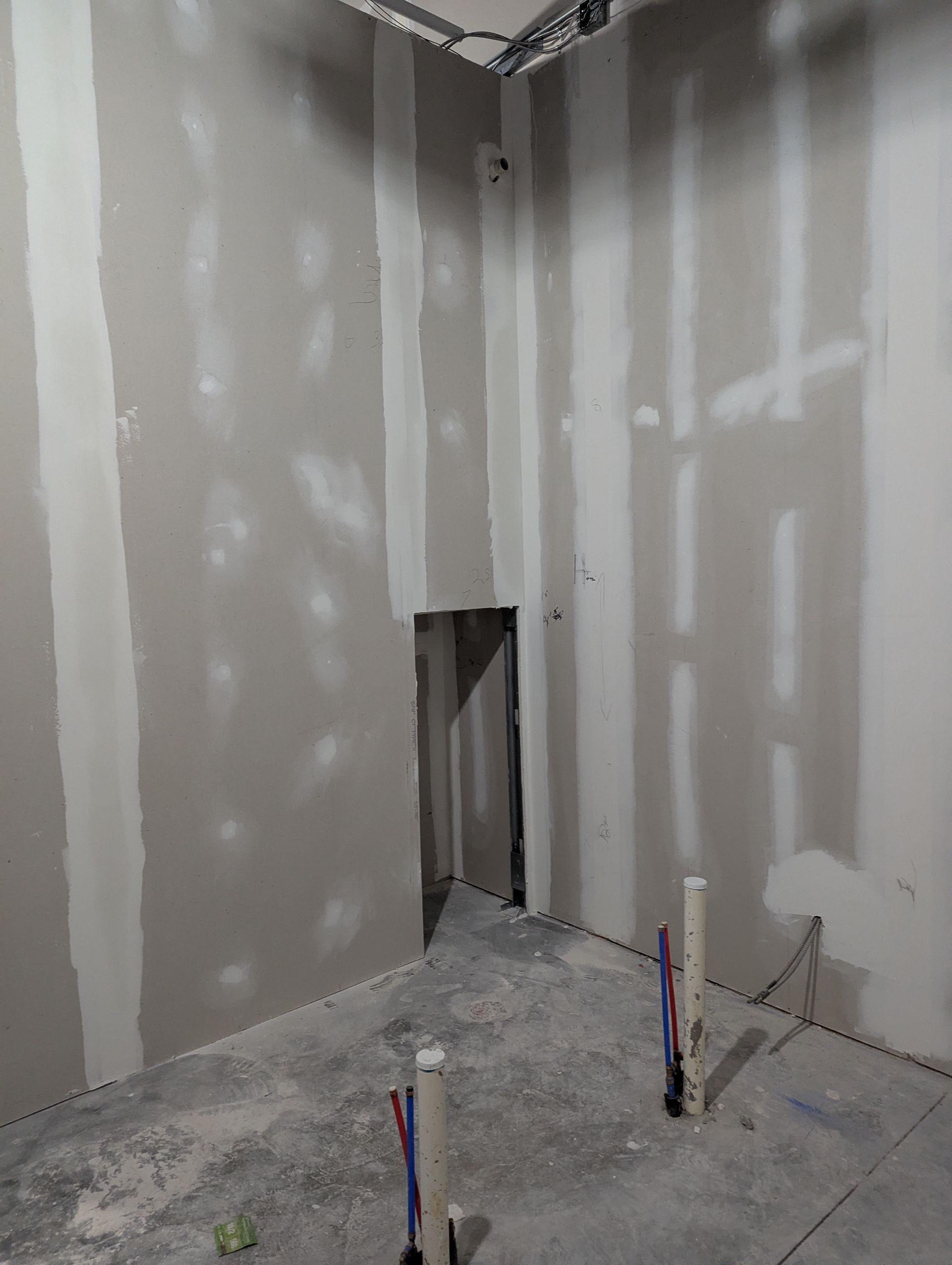 Unfinished interior room with drywall, taped seams, a wall cutout, and exposed utility pipes sticking out of the floor.