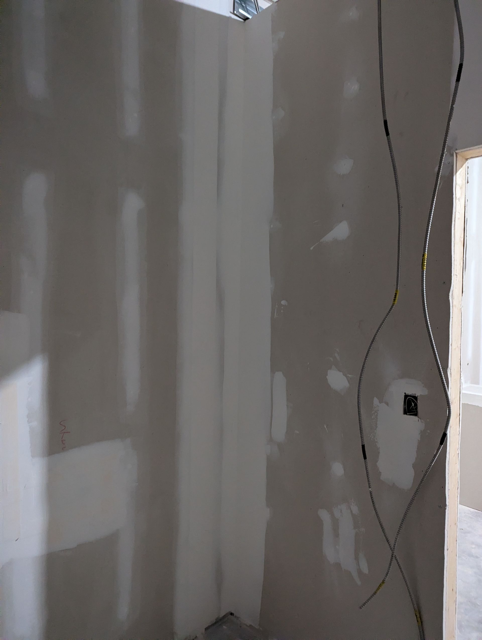 Interior view of an unfinished wall with taped drywall joints and exposed electrical wires hanging down.