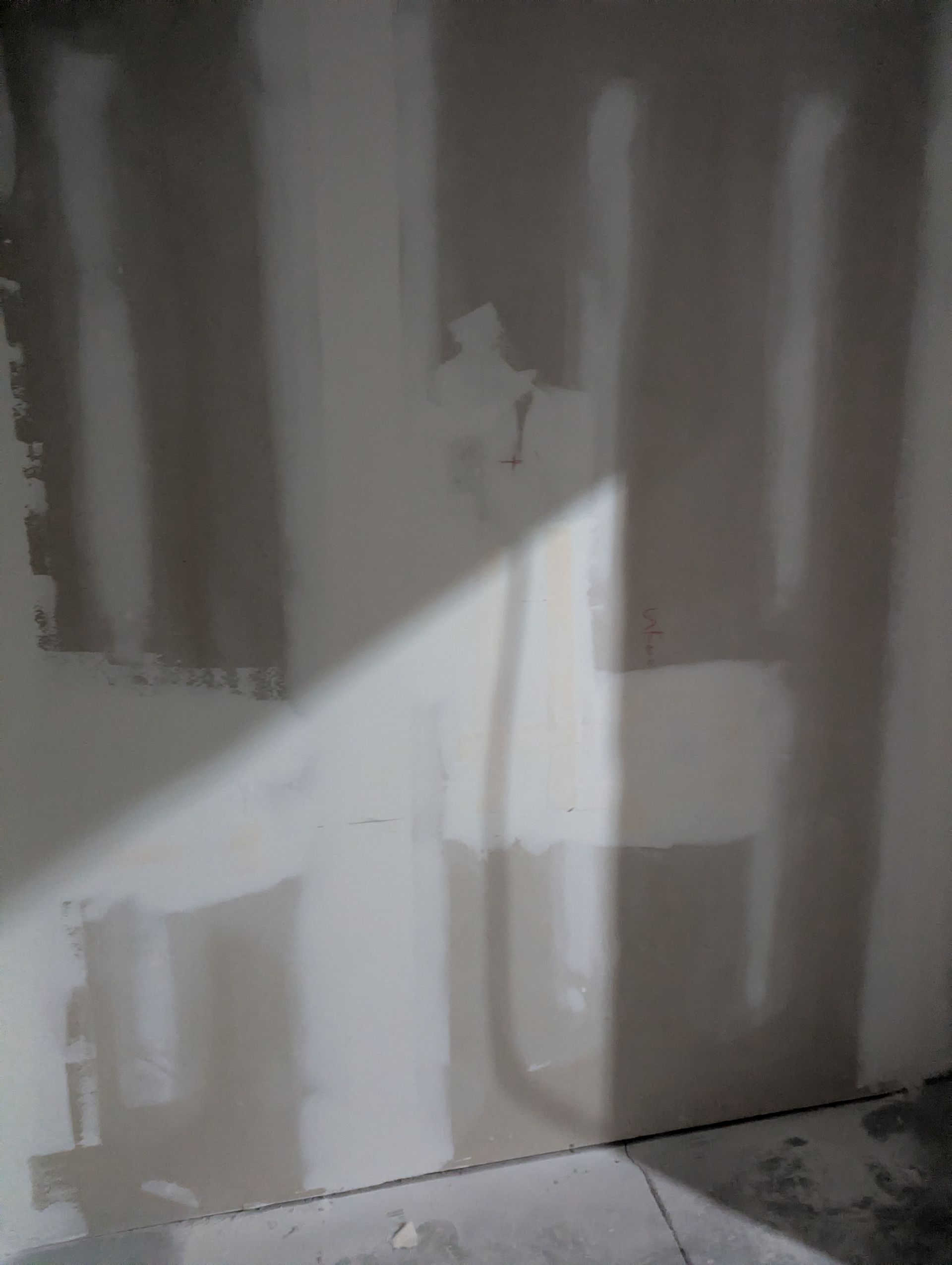 A light-colored interior wall with drywall seams covered in patching compound, illuminated by a diagonal beam of sunlight.