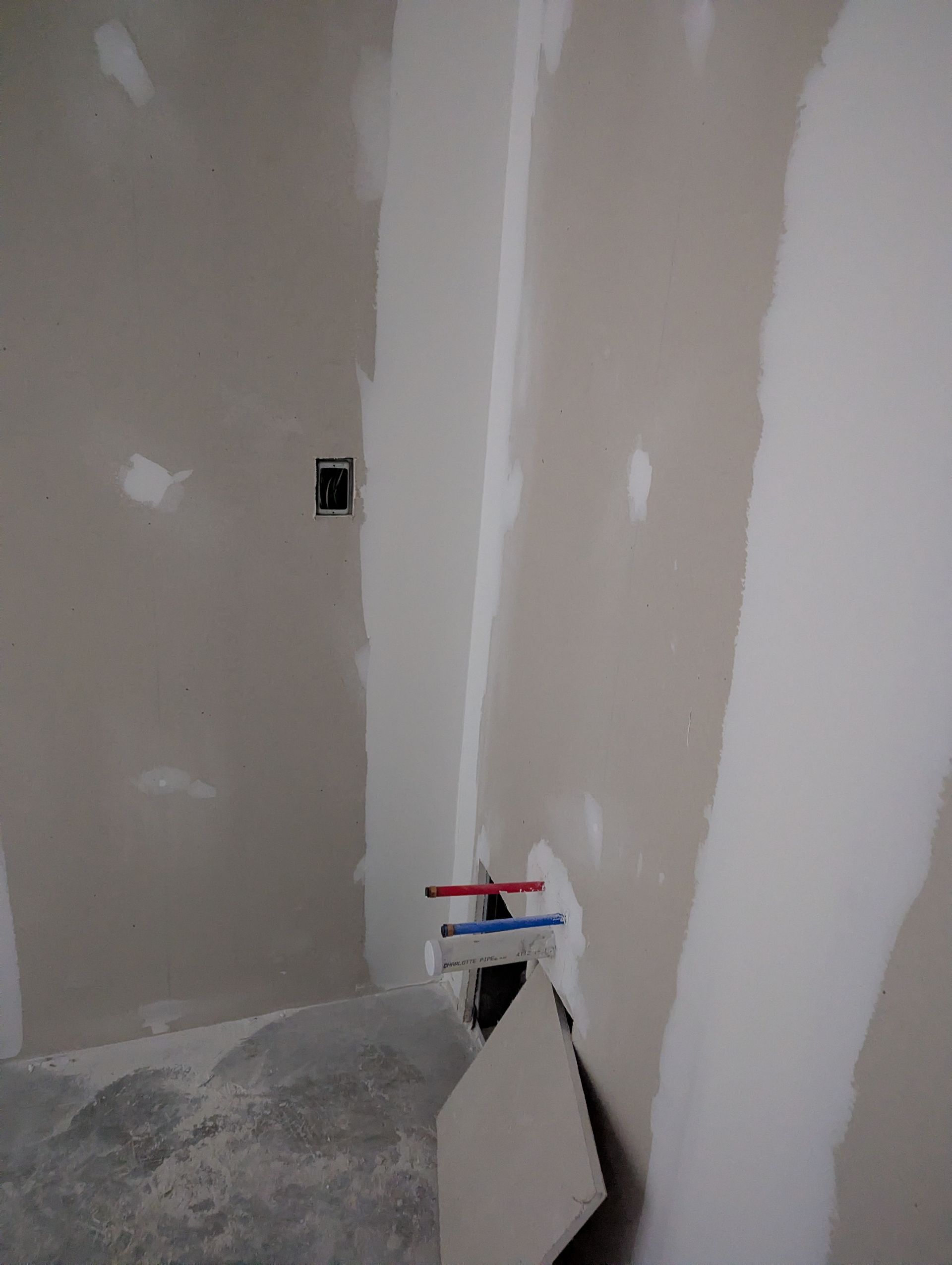 An unfinished interior wall with drywall seams, an electrical outlet, and red and blue plumbing pipes protruding from below.