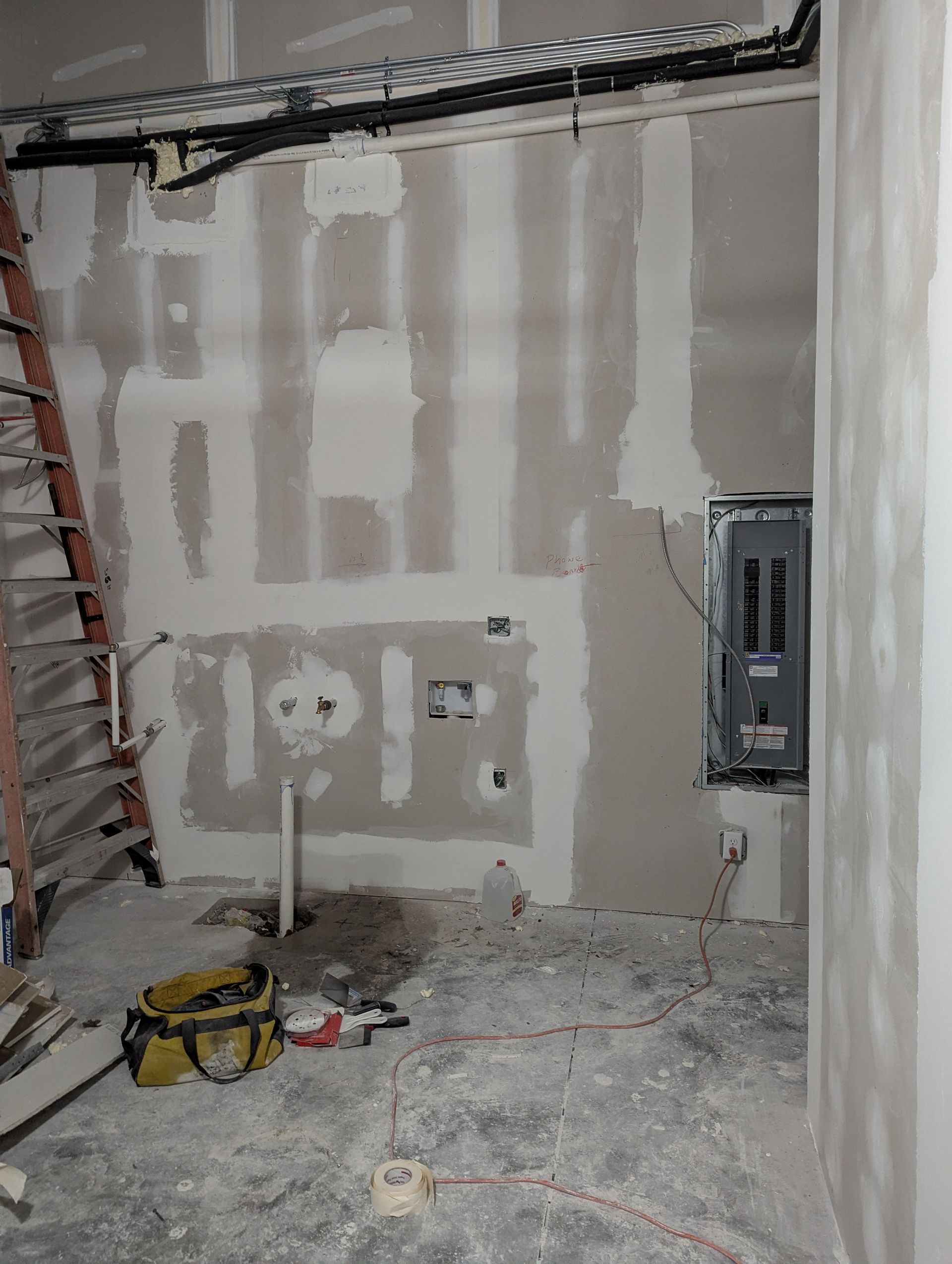 An unfinished room under construction with drywall patches, a ladder, a central electrical panel, and exposed wiring.