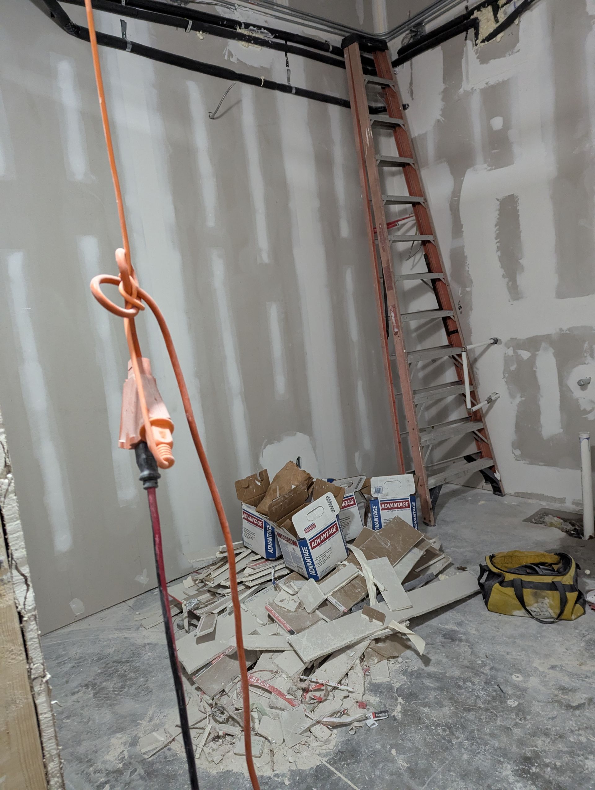 An unfinished room under construction, with a ladder leaned against a wall, debris on the floor, and hanging electrical wire.
