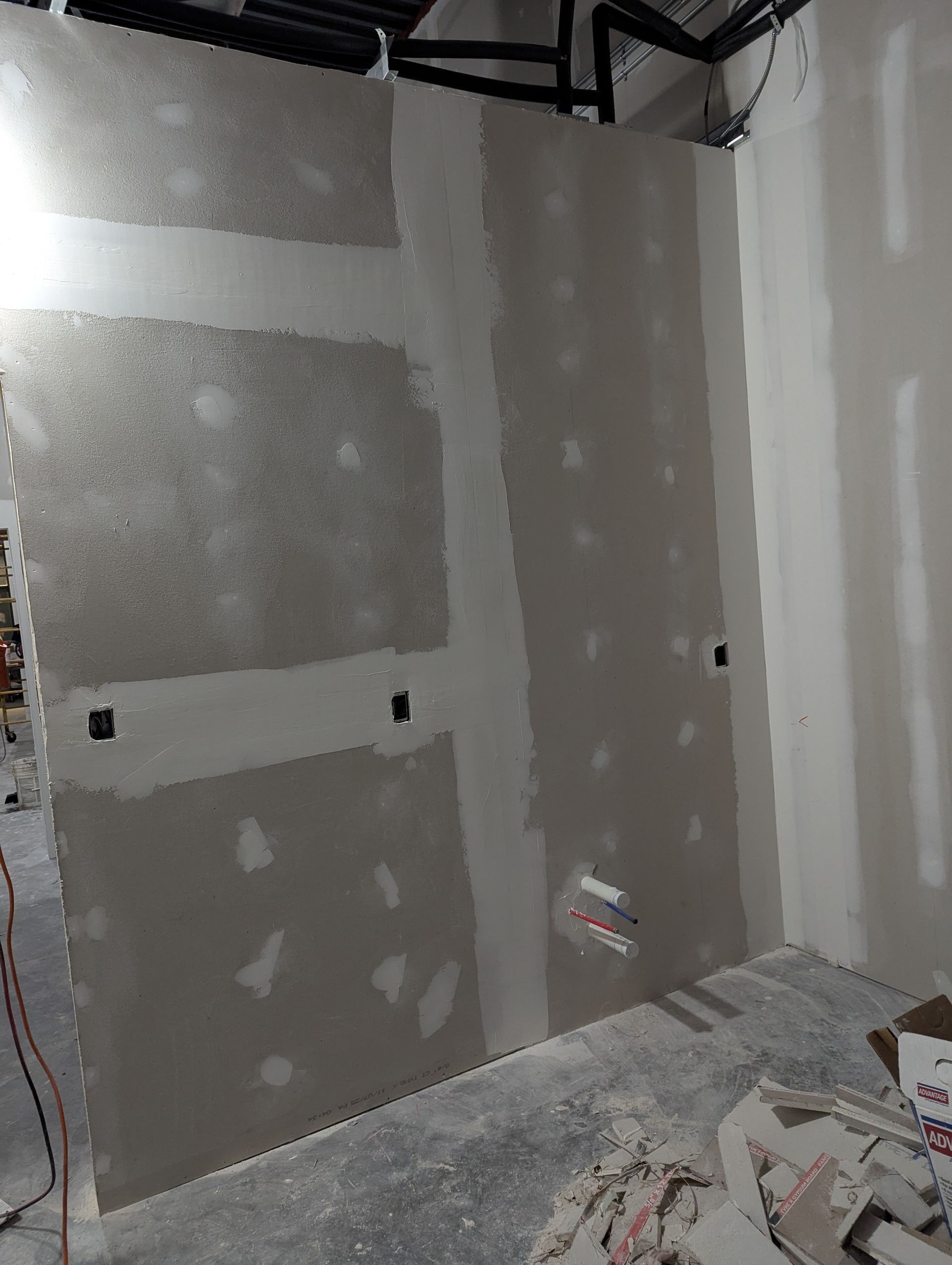 A partially finished drywall wall with taped seams, electrical outlet boxes, and a plumbing rough-in in a construction site.