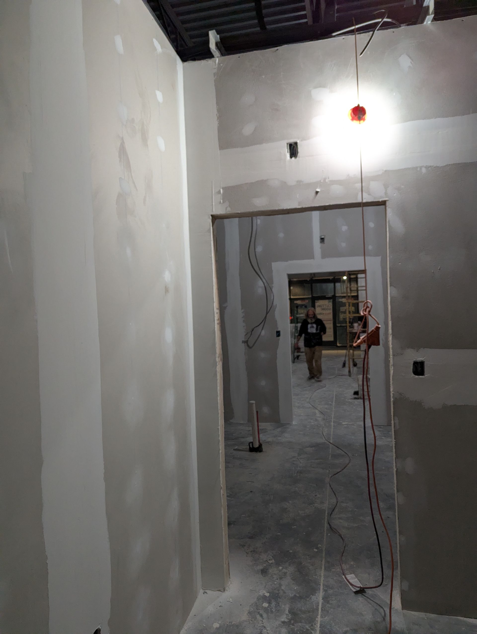 A narrow hallway under construction with unfinished drywall, a single hanging light, and a person in the distance.