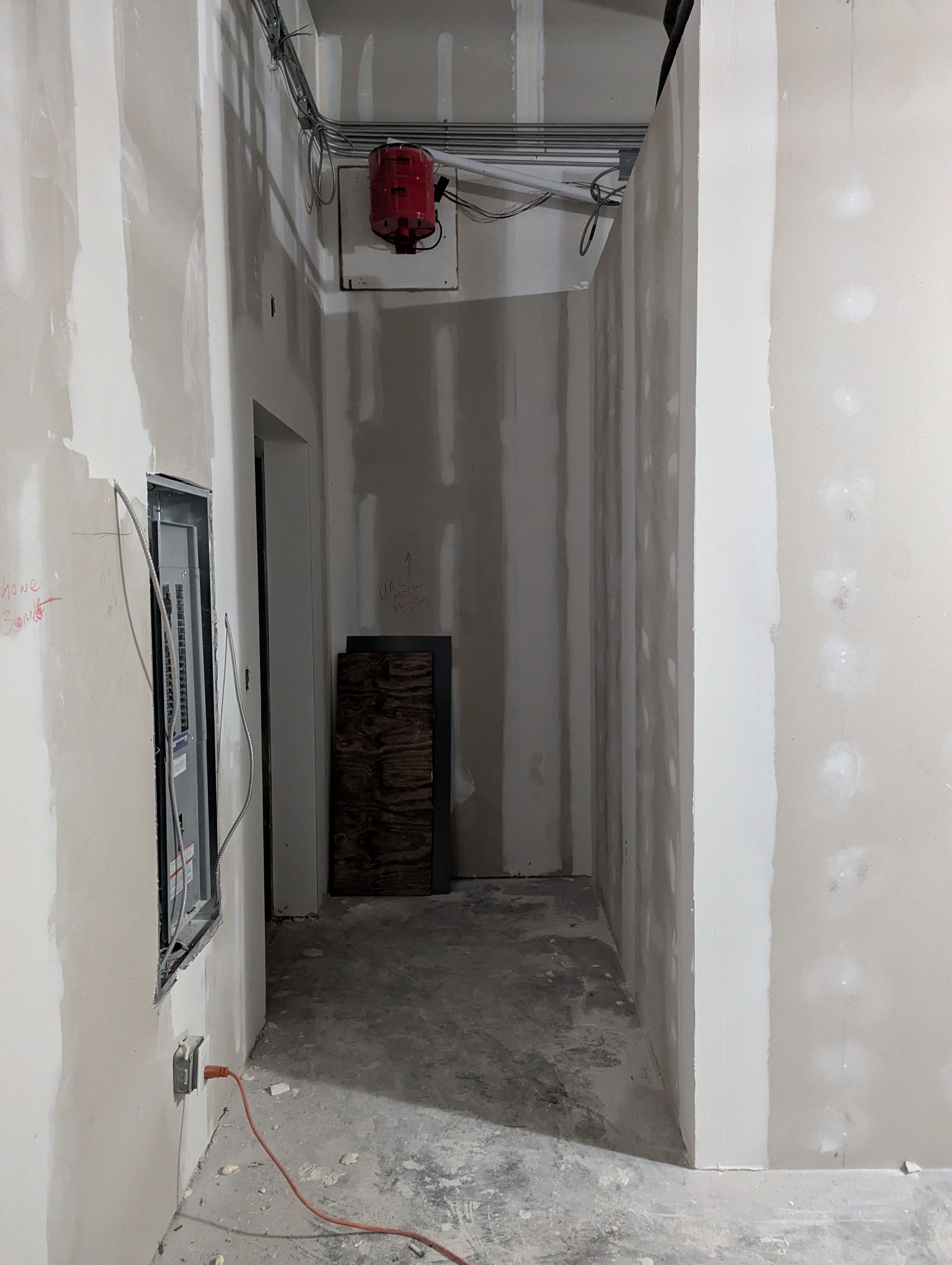 An unfinished construction site showing an interior hallway with unpainted drywall, exposed ceiling wires, and a breaker box.