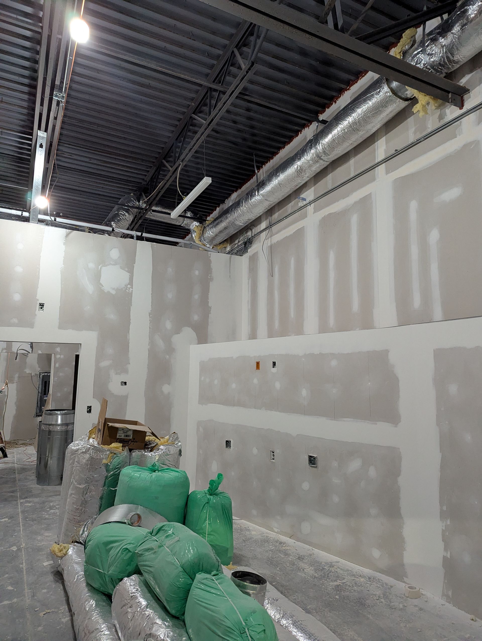 An unfinished interior room with drywall, exposed ceiling joists, metal ductwork, and green bags of construction materials.