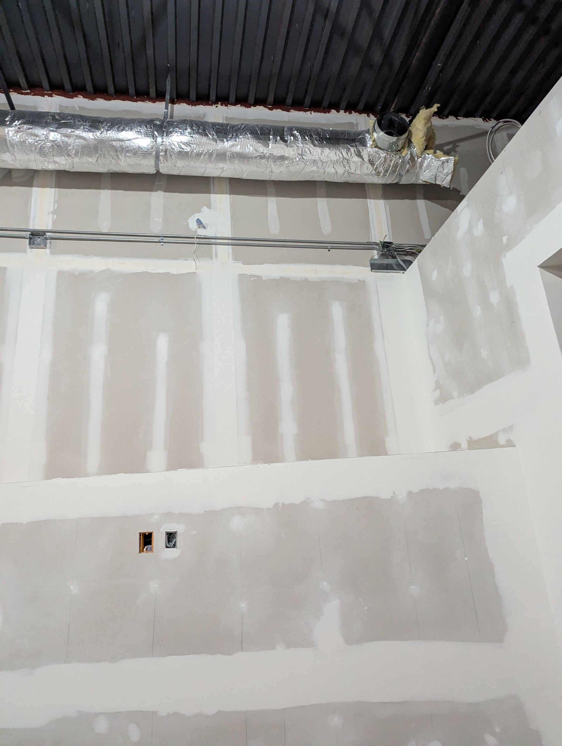 Unfinished drywall installation on walls and an exposed corrugated ceiling with an insulated air duct and electrical work.
