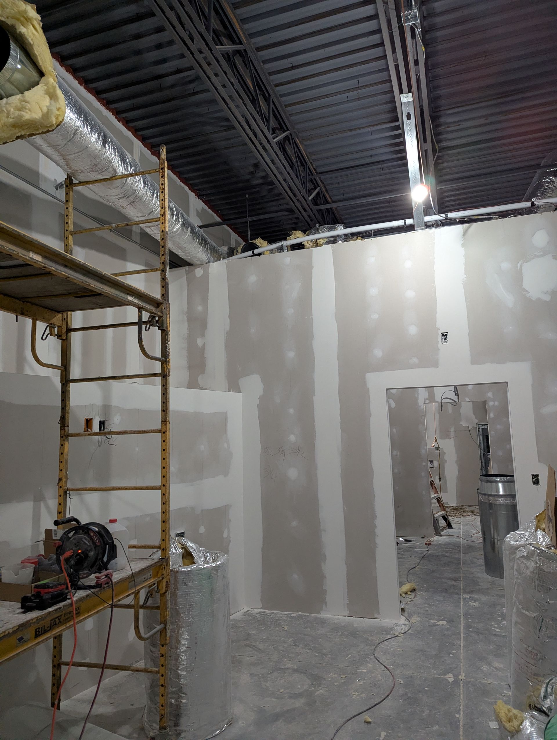 Interior construction site featuring drywall panels on metal studs, a tall scaffolding tower, and an open doorway.