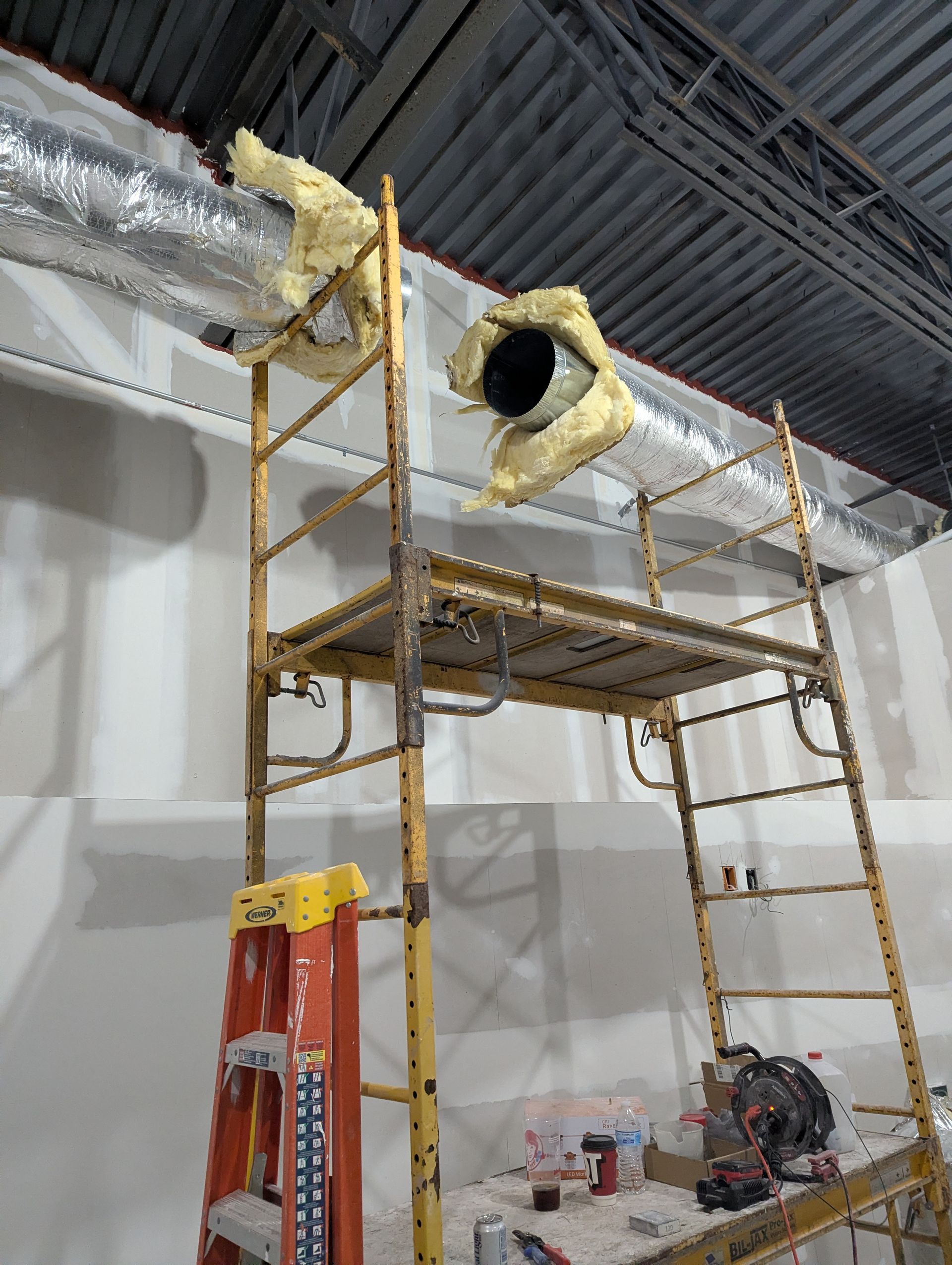 A yellow metal scaffolding unit stands in a construction zone, with foam sealant around HVAC ducts near the ceiling.