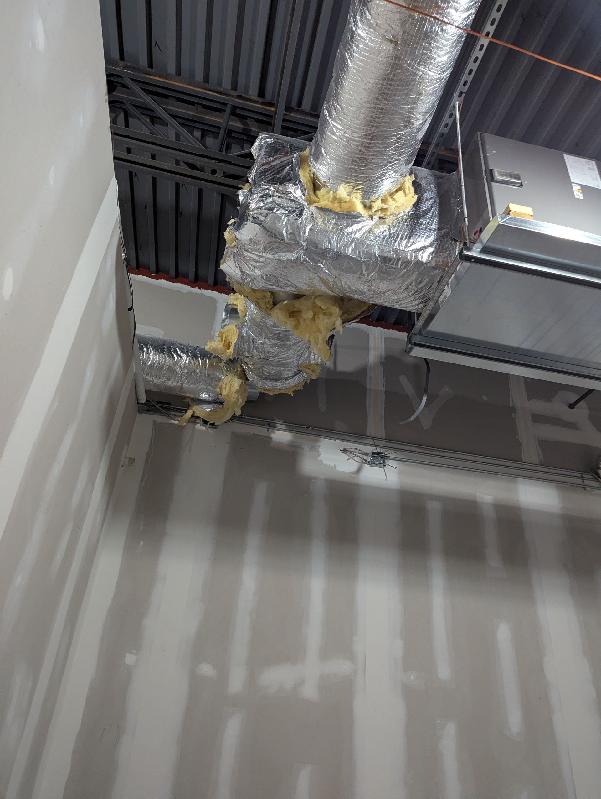 Exposed HVAC ductwork with yellow spray foam insulation gaps, installed near a corner in a room with unfinished drywall.