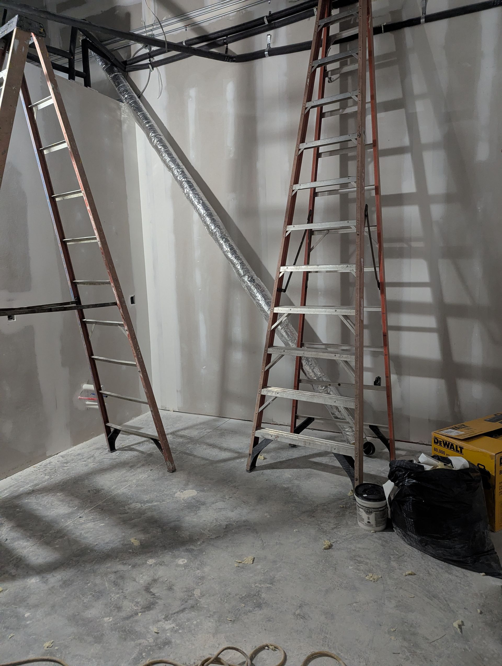 Two tall stepladders stand on a concrete floor in an unfinished room with exposed pipes and drywall.